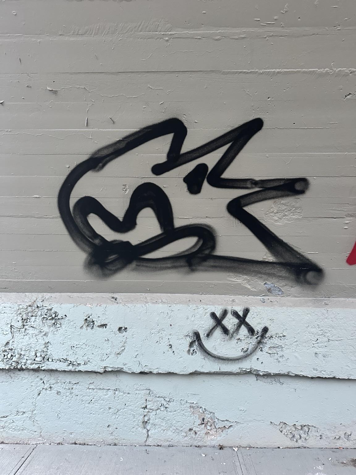 a black outline of sonic the hesgehog's spraypainted on a tan and white wall. a smiley face with Xs for eyes is below to the right