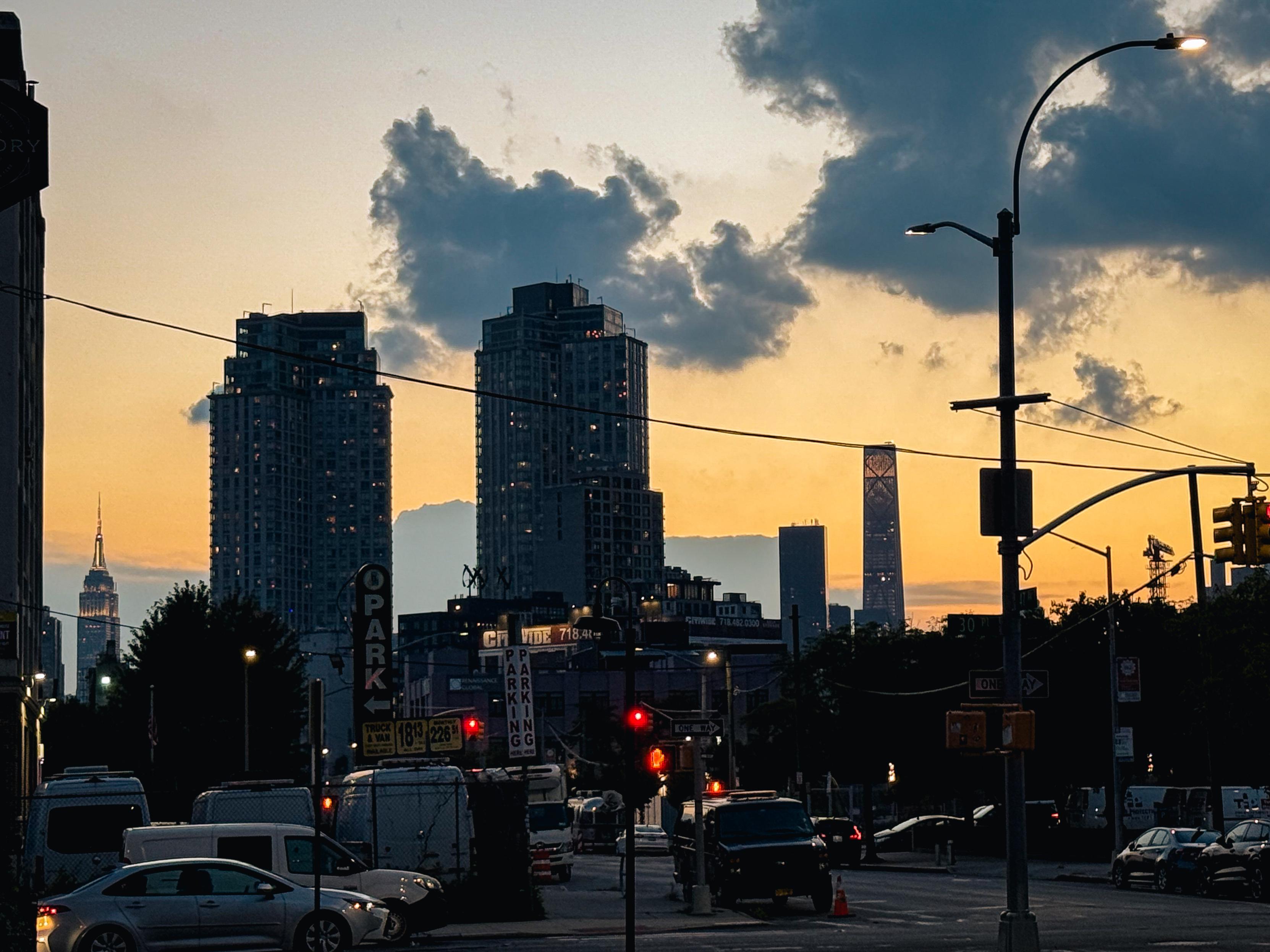 Part of the Long Island City skyline at sunset featuring tall buildings, including the Empire State Building on the left in nearby Manhattan. Streetlights and traffic signals are visible in the foreground, with a colorful sky blending orange and blue hues, scattered clouds present.
