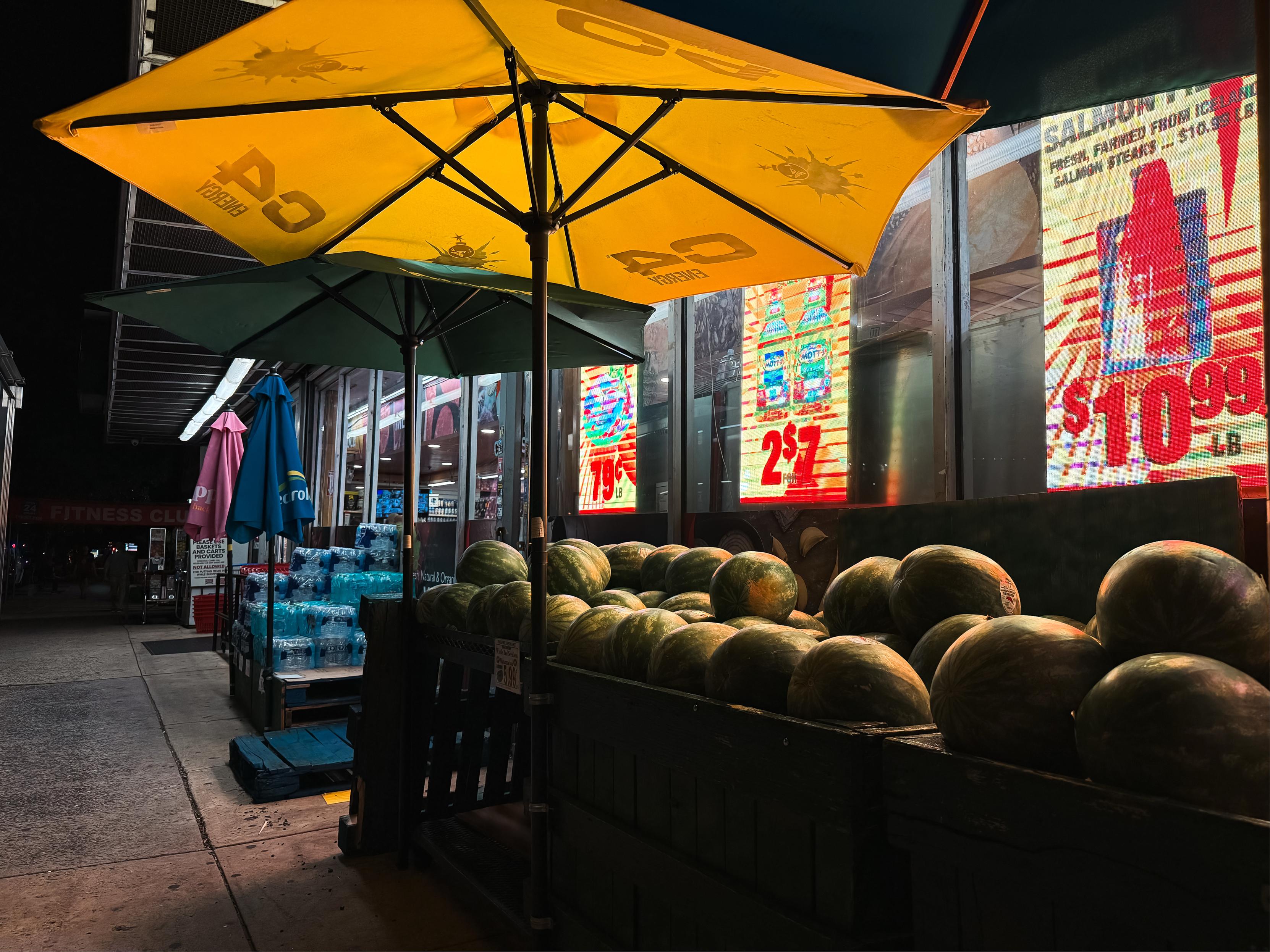 A nighttime scene featuring a display of watermelons under yellow and green umbrellas outside a supermarket. In the background, colorful neon signs advertise prices for various items, including bottled drinks. Stacked packs of bottled water are visible on the side.