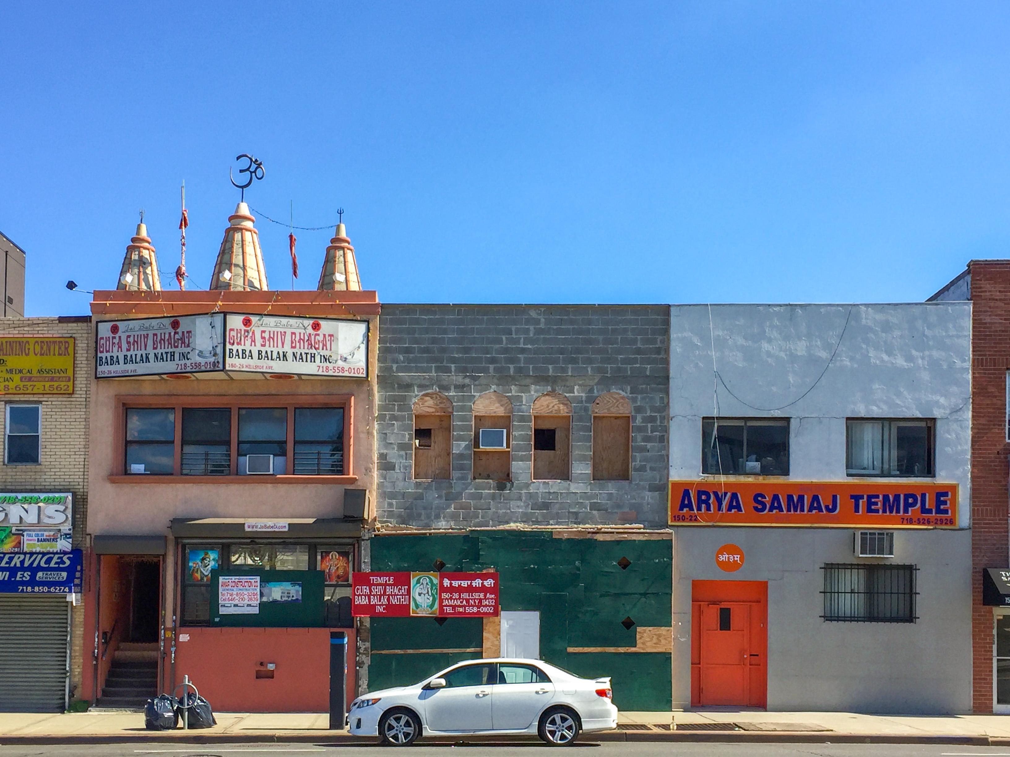 A street scene in Jamaica, Queens. A long white sedan is parked in front of two South Asian religious temples.