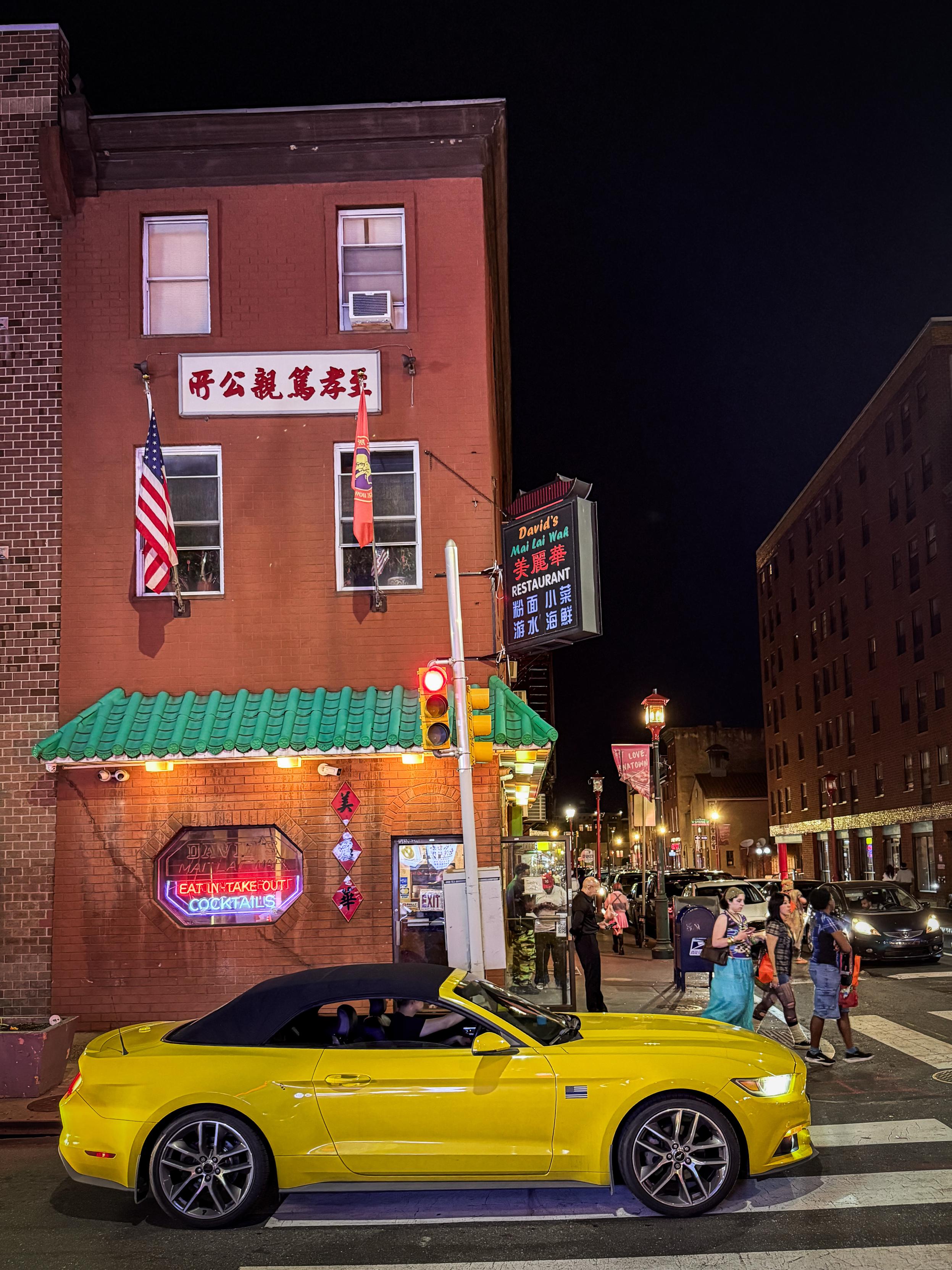 A yellow sports car sits at a red light in front of a Chinese restaurant on the corner. Pedestrians walk by through the crosswalk. It is night time and vibrant lights from businesses along with streetlights help to illuminate the scene.