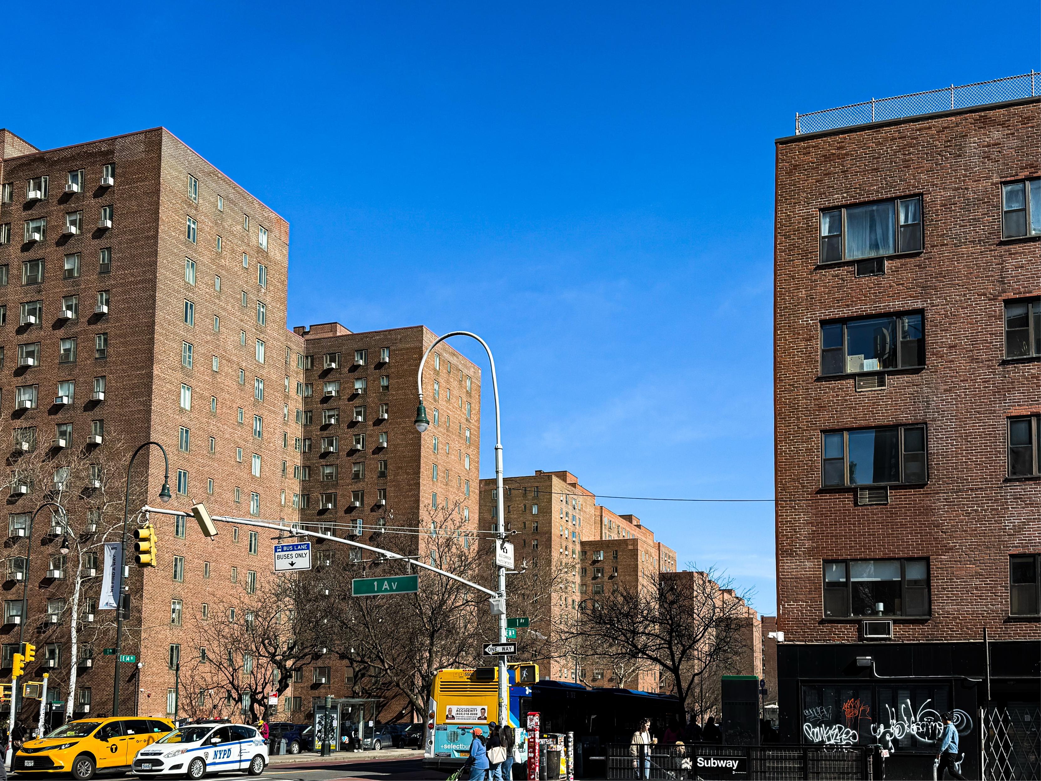 A streetscape featuring red brick apartment buildings under a clear blue sky. There are yellow taxis and a bus stop, along with street signage indicating traffic regulations. A subway entrance is visible on the left.