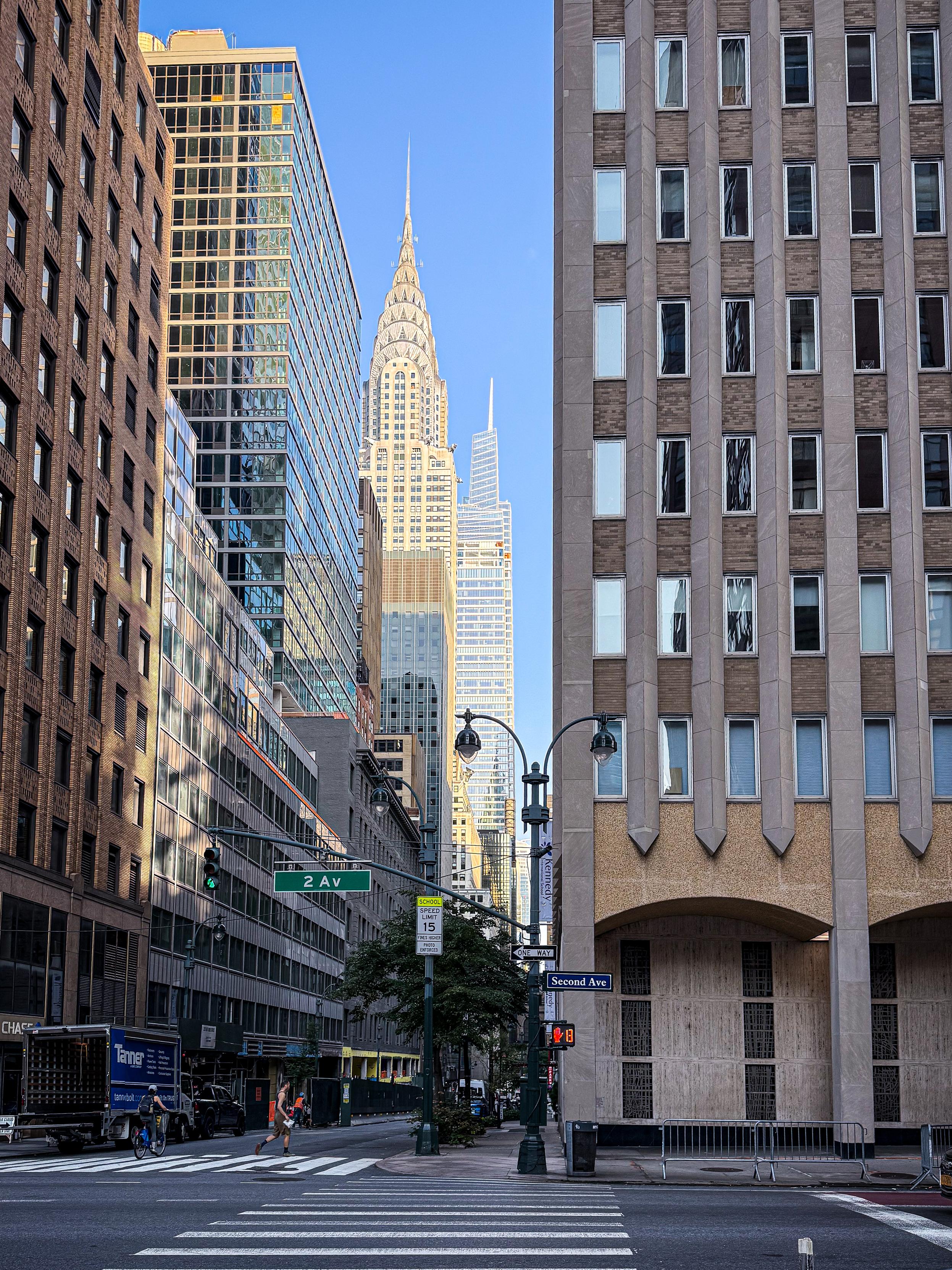 A view of a Manhattan city street with skyscrapers, including the Chrysler Building, prominently visible. The scene features a traffic signal, street signs for “2 Av” and “Second Ave,” and a pedestrian crossing.