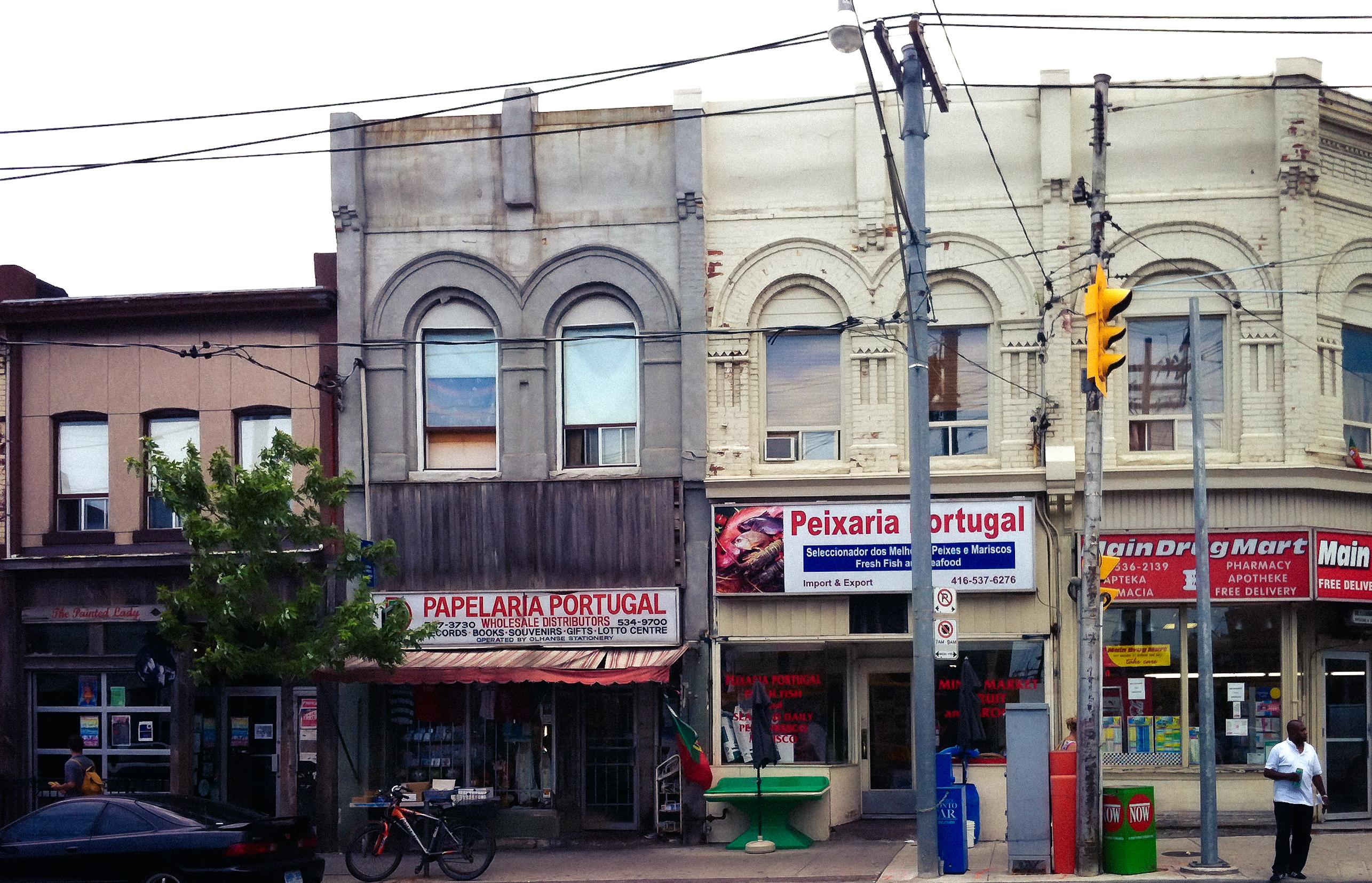 An urban Toronto street scene featuring several small businesses. The left side displays a shopfront labeled "PAPELARIA PORTUGAL" with a striped awning. Adjacent to it is a store named "Peixaria Portugal," which specializes in seafood.