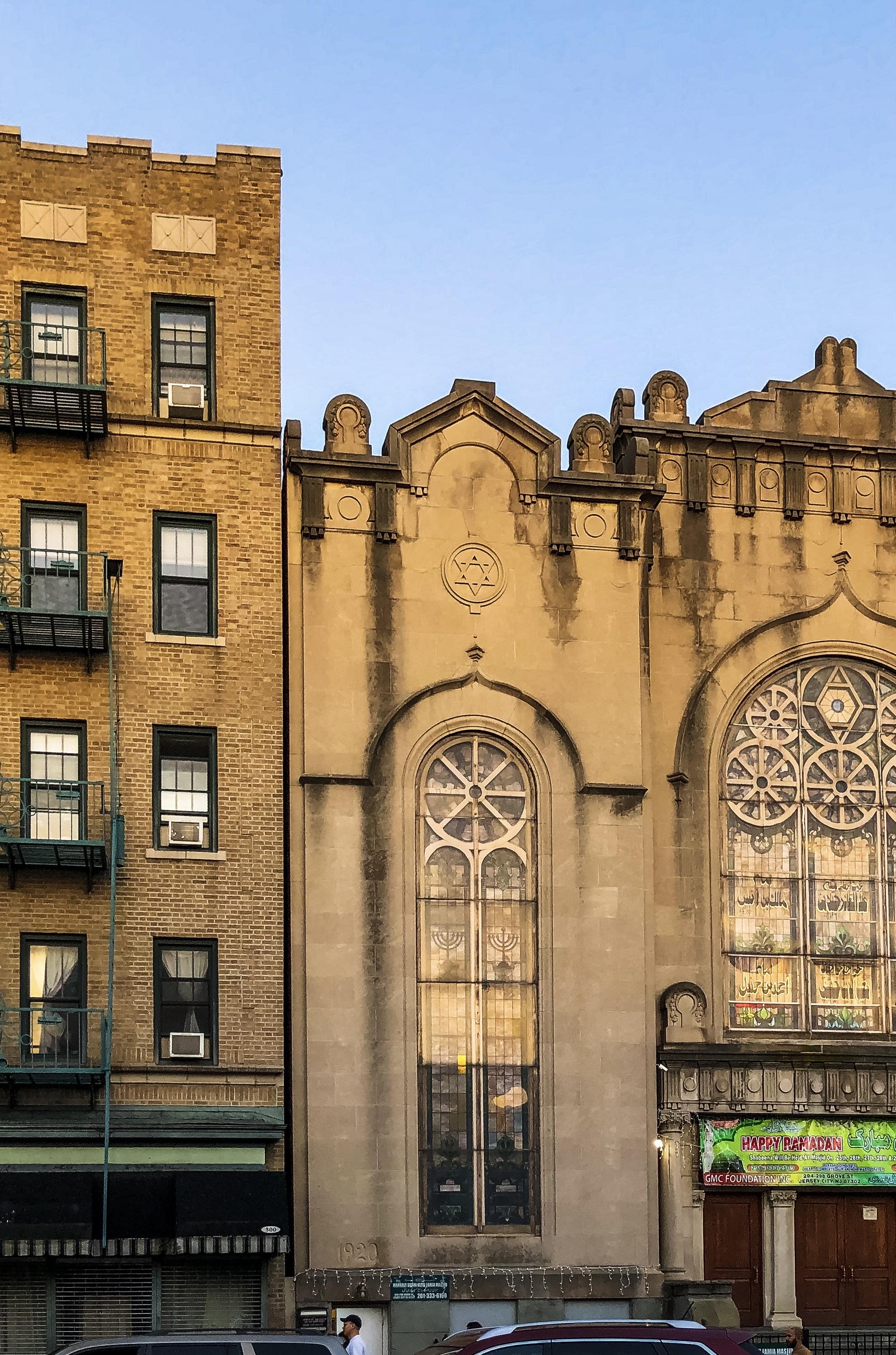 The image shows a historic building serving as a mosque featuring a large arched window with stained glass. Next to it is an apartment building with a brick facade and fire escapes. A banner on the building indicates a celebration or event related to Ramadan. The setting is urban.