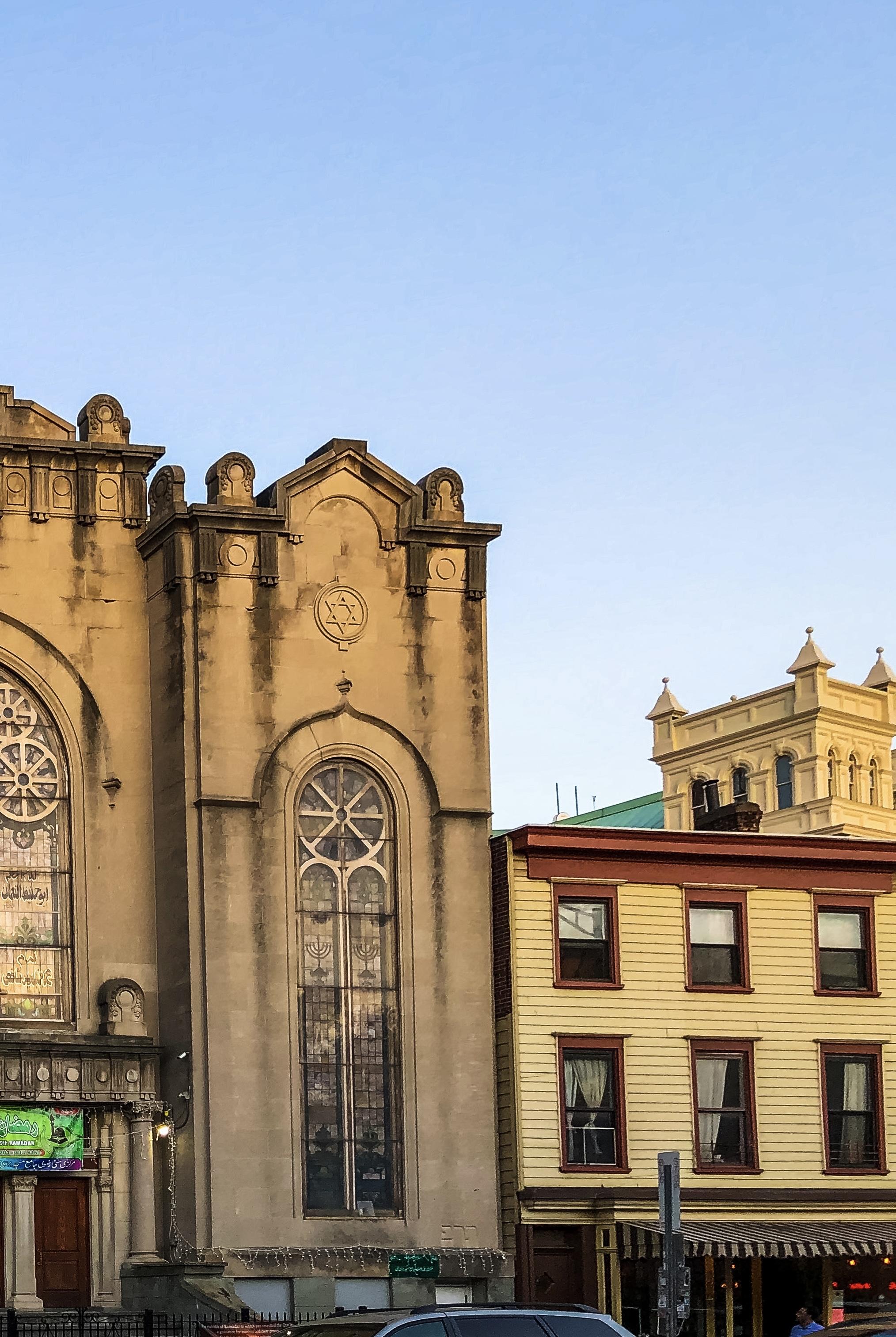 Image features a large historic building serving as a mosque with decorative architecture and prominent stained glass windows on the left. Adjacent to it is a contrasting yellow and red building with a striped awning. The sky is clear and blue.