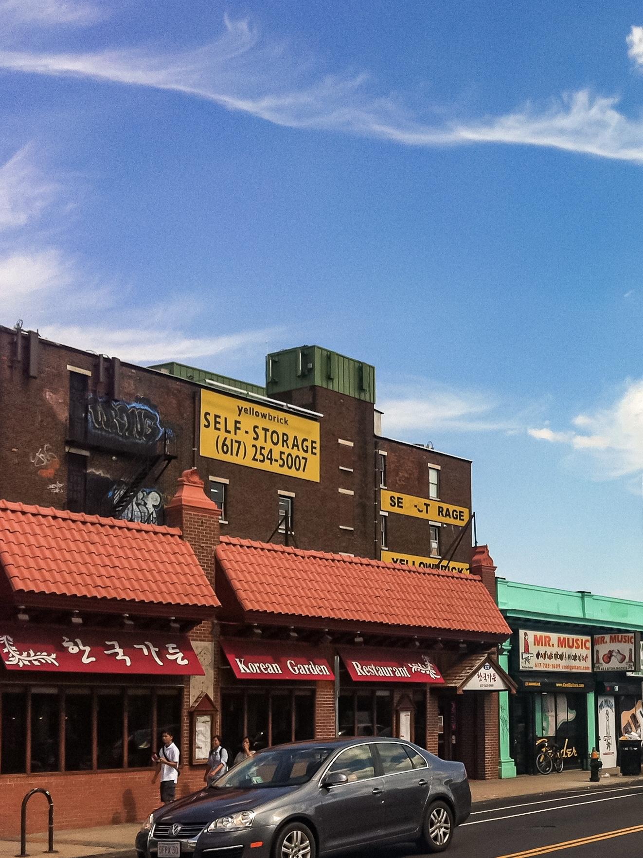 An Allston street scene featuring several buildings. The foreground includes a Korean restaurant with red signage. Behind it is a building with a yellow "Self Storage" sign. The sky is clear blue with wispy clouds, and there's a gray car parked on the street.