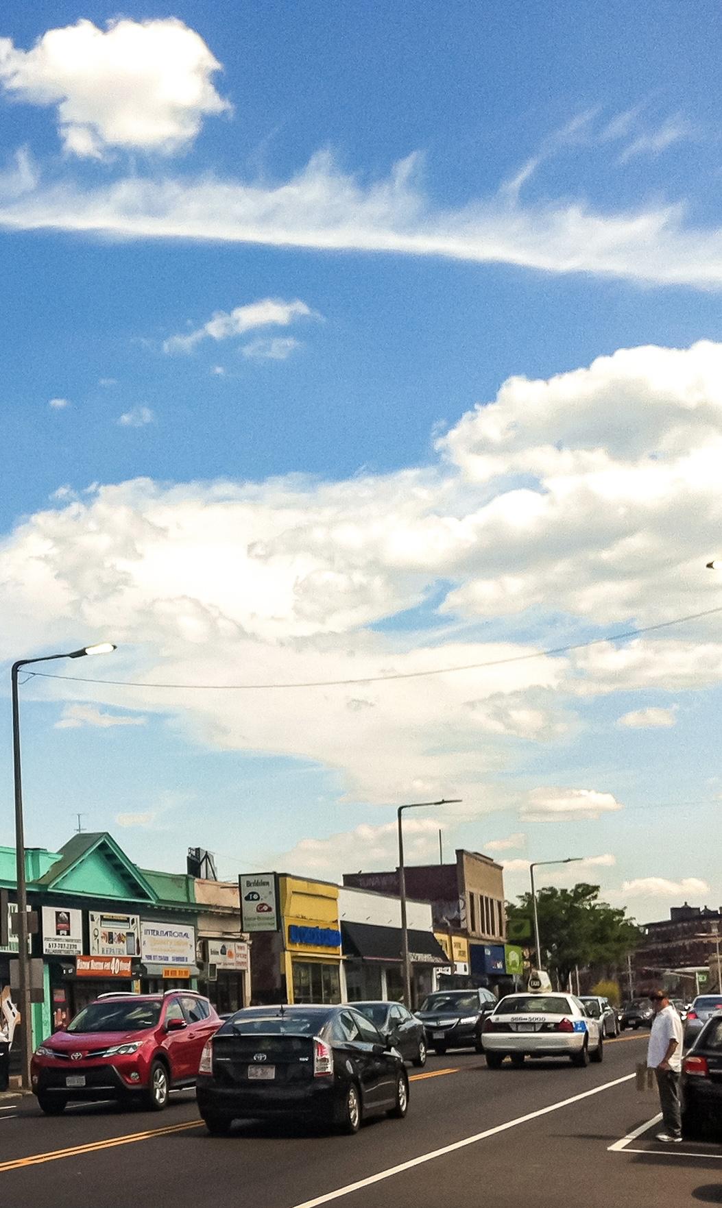 A busy Allston street scene featuring several cars, including a taxi, on the road. There are shops on either side with various signs, under a blue sky with scattered clouds. A pedestrian is standing on the sidewalk.