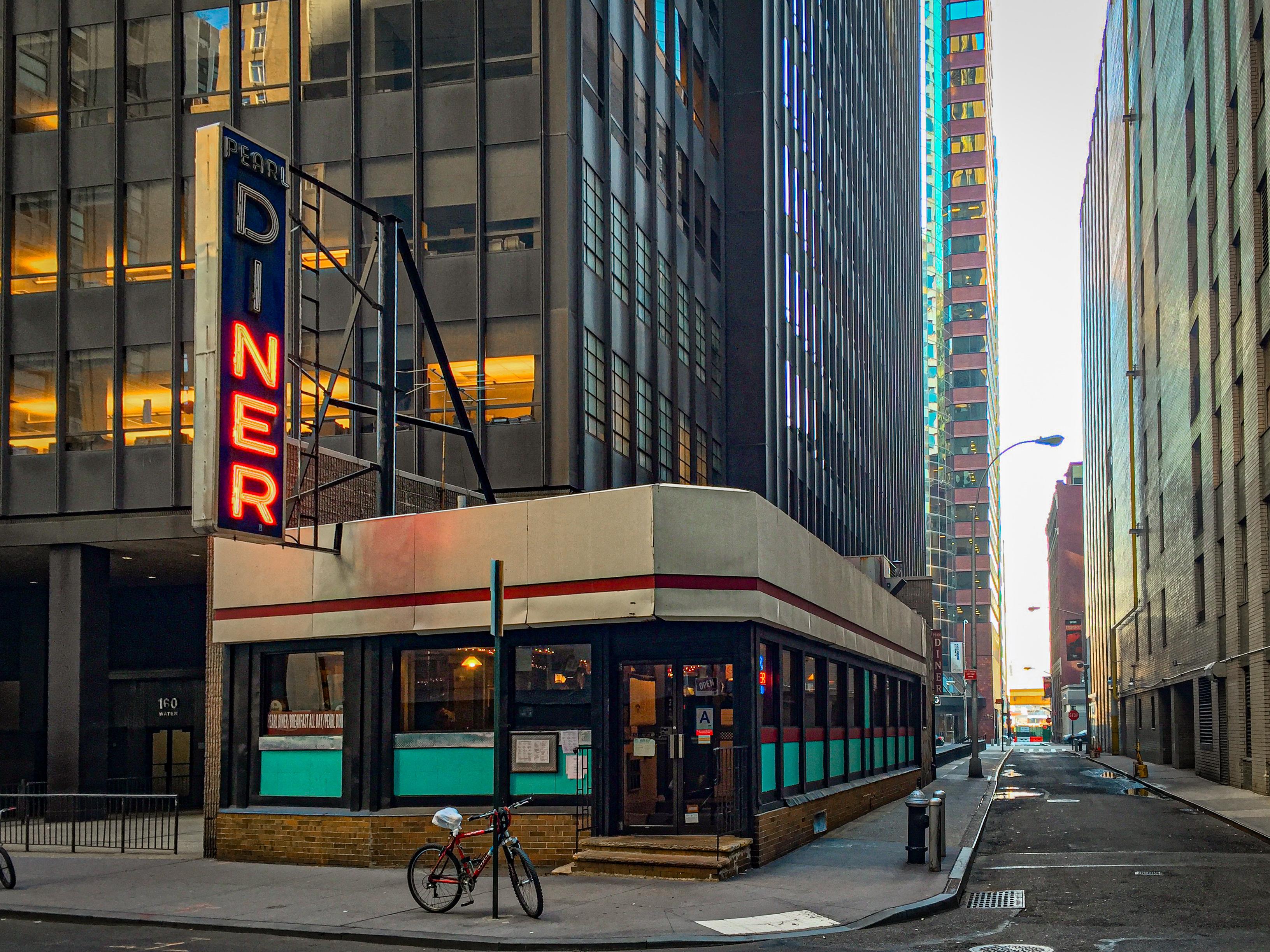 Image of a street scene in lower Manhattan featuring a diner with a bright neon sign that reads "PEARL DINER." The diner has a modern yet retro design, located between tall, glass-sided buildings. A bicycle is parked nearby, and the street appears quiet.