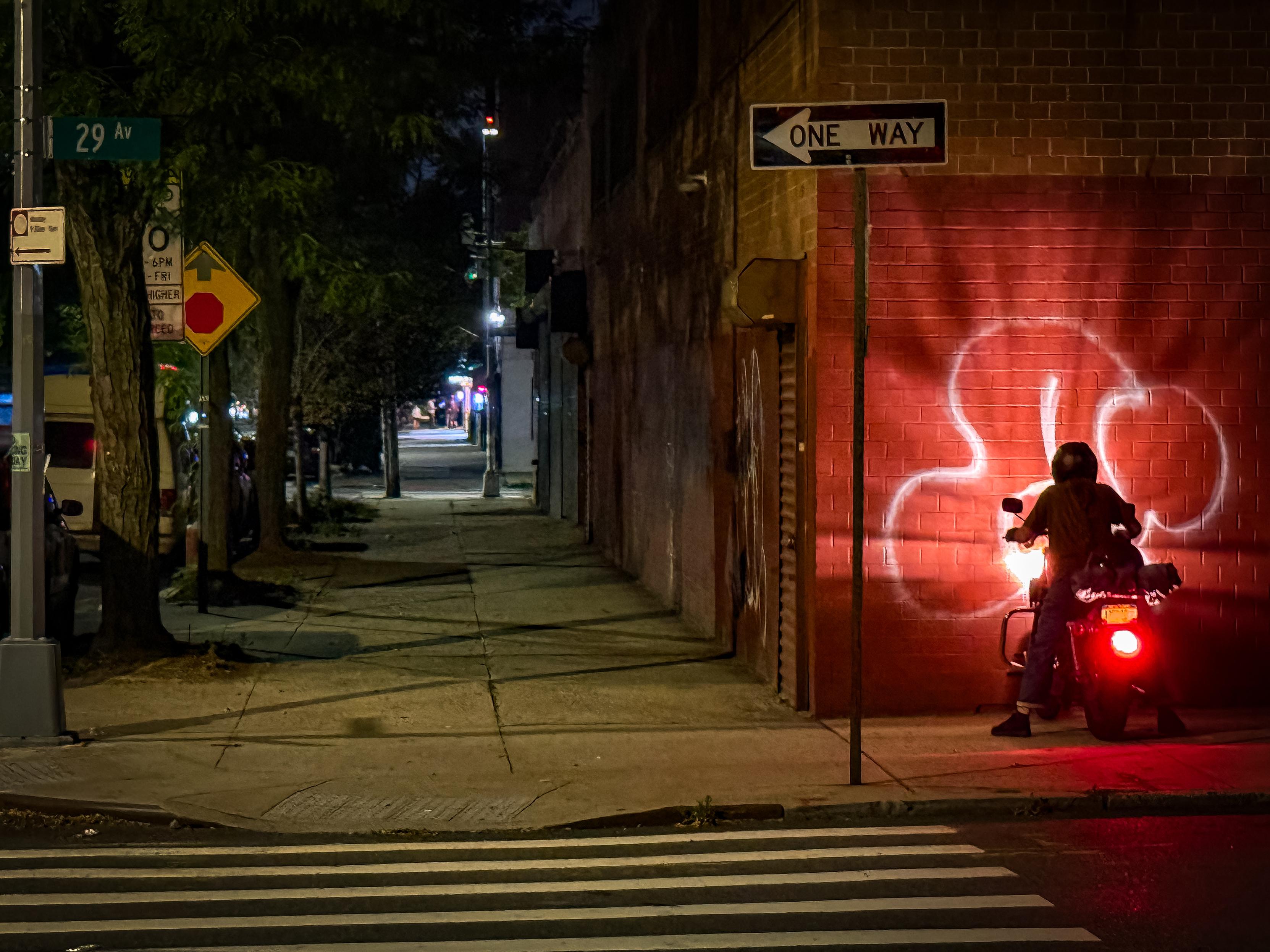 A dimly lit Astoria, Queens street at night featuring a motorcycle revving its engine beside a brick wall with its lights illuminating the area. The wall has a graffiti design, and a street sign indicates "One Way." Trees line the sidewalk, and various urban elements are visible in the background.