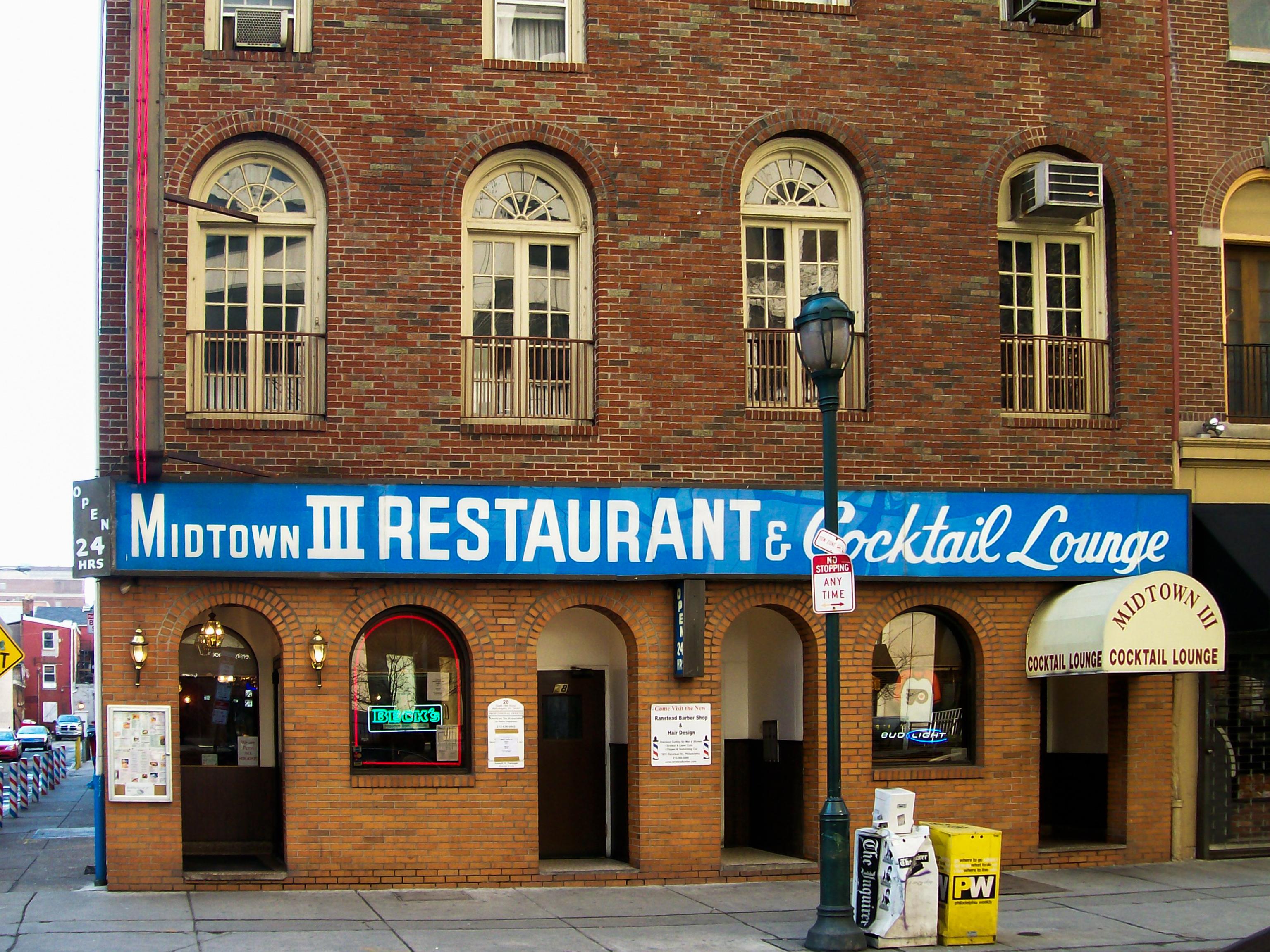 A brick building featuring a sign for "Midtown III Restaurant & Cocktail Lounge." The facade includes arched windows and two entrances, with light fixtures and a yellow newspaper box in front. A sign indicates it is open 24 hours.