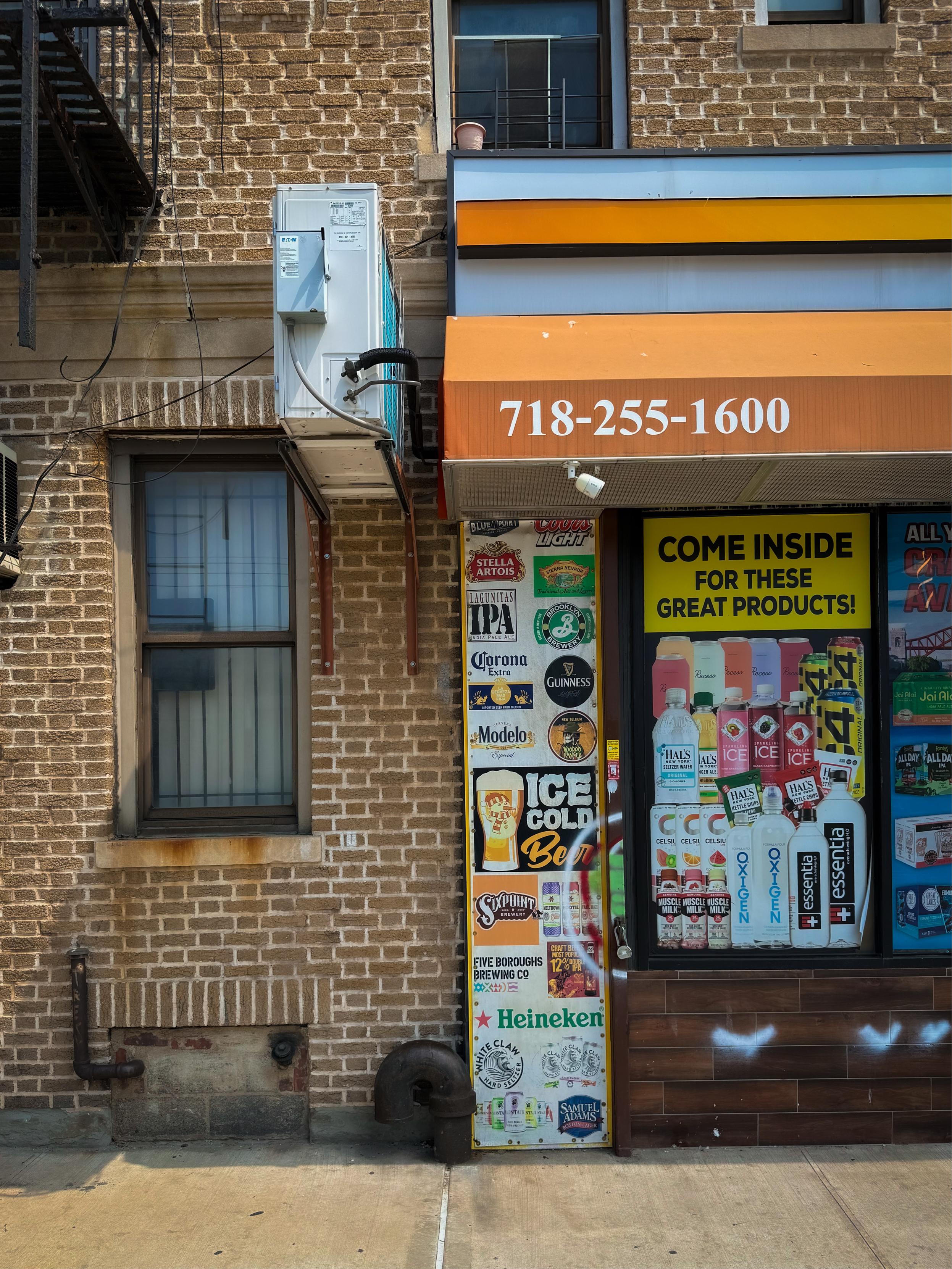 The image shows a brick building with a deli storefront featuring a colorful orange-hued awning displaying a phone number. The entrance has a variety of beverage advertisements, including beers and energy drinks. An air conditioning unit is mounted above a window in a neighboring residential unit.