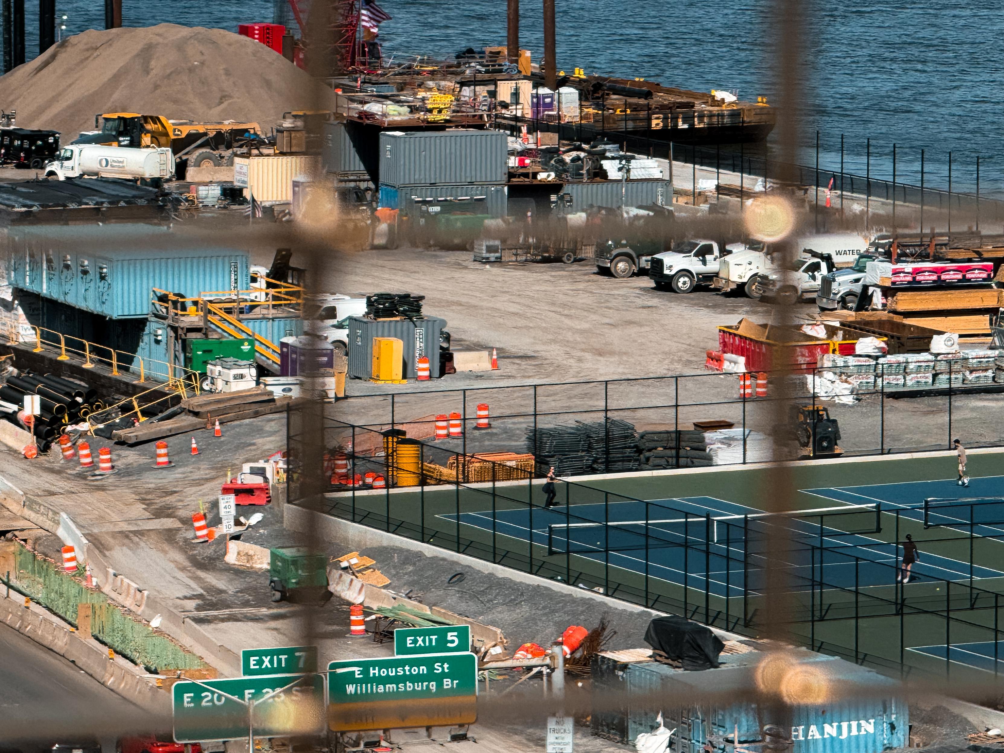 A construction site near the East River, featuring various machinery and shipping containers. In the foreground, there are two tennis courts with players, and traffic signs indicating nearby exits. The scene is framed by a fence along the Williamsburg Bridge.