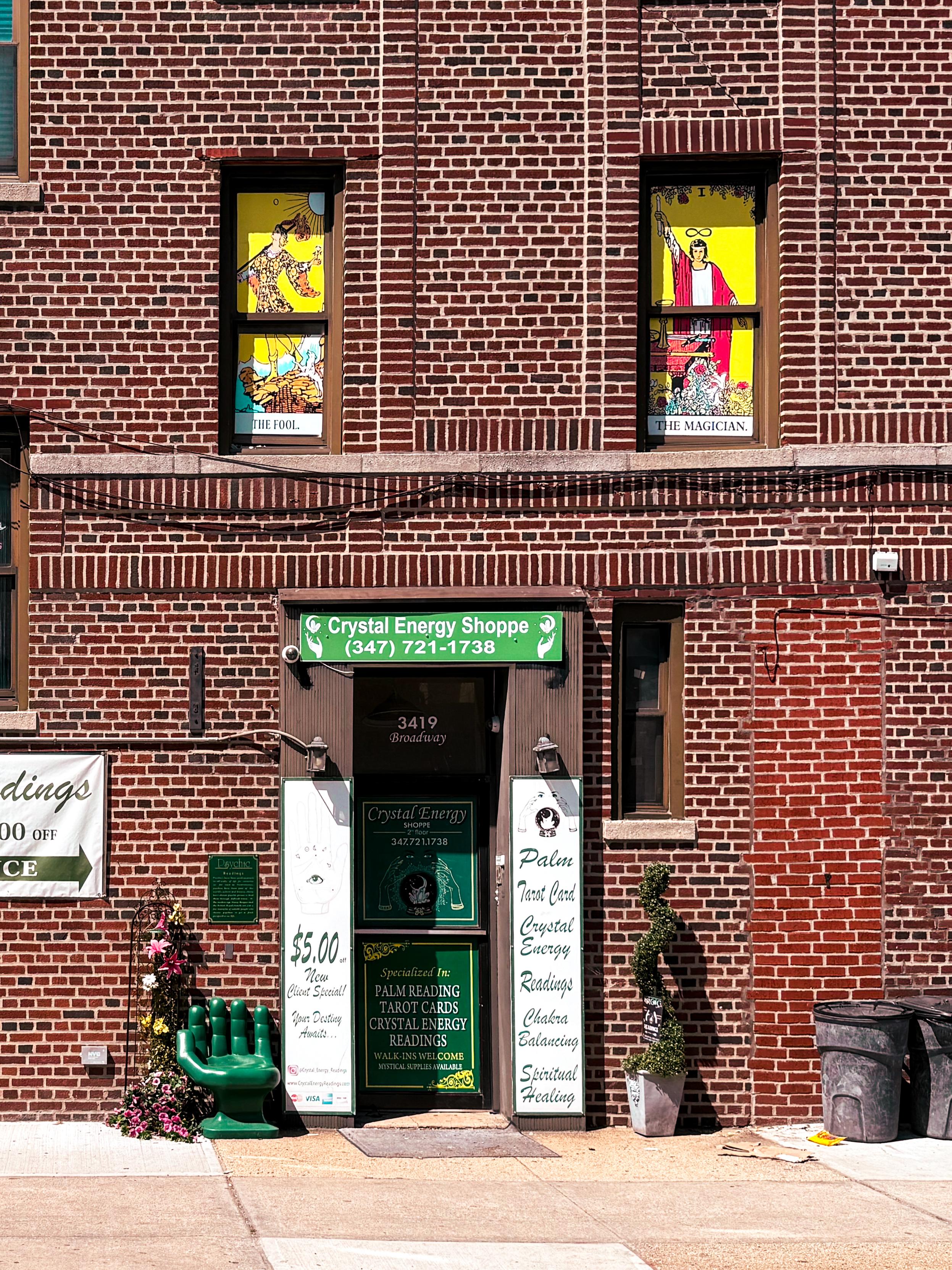 The image shows a brick building with a sign for the "Crystal Energy Shoppe" located at 3419 Broadway. The entrance features green signage and promotions for palm readings, tarot cards, and crystal energy readings. Two windows display colorful tarot card illustrations.