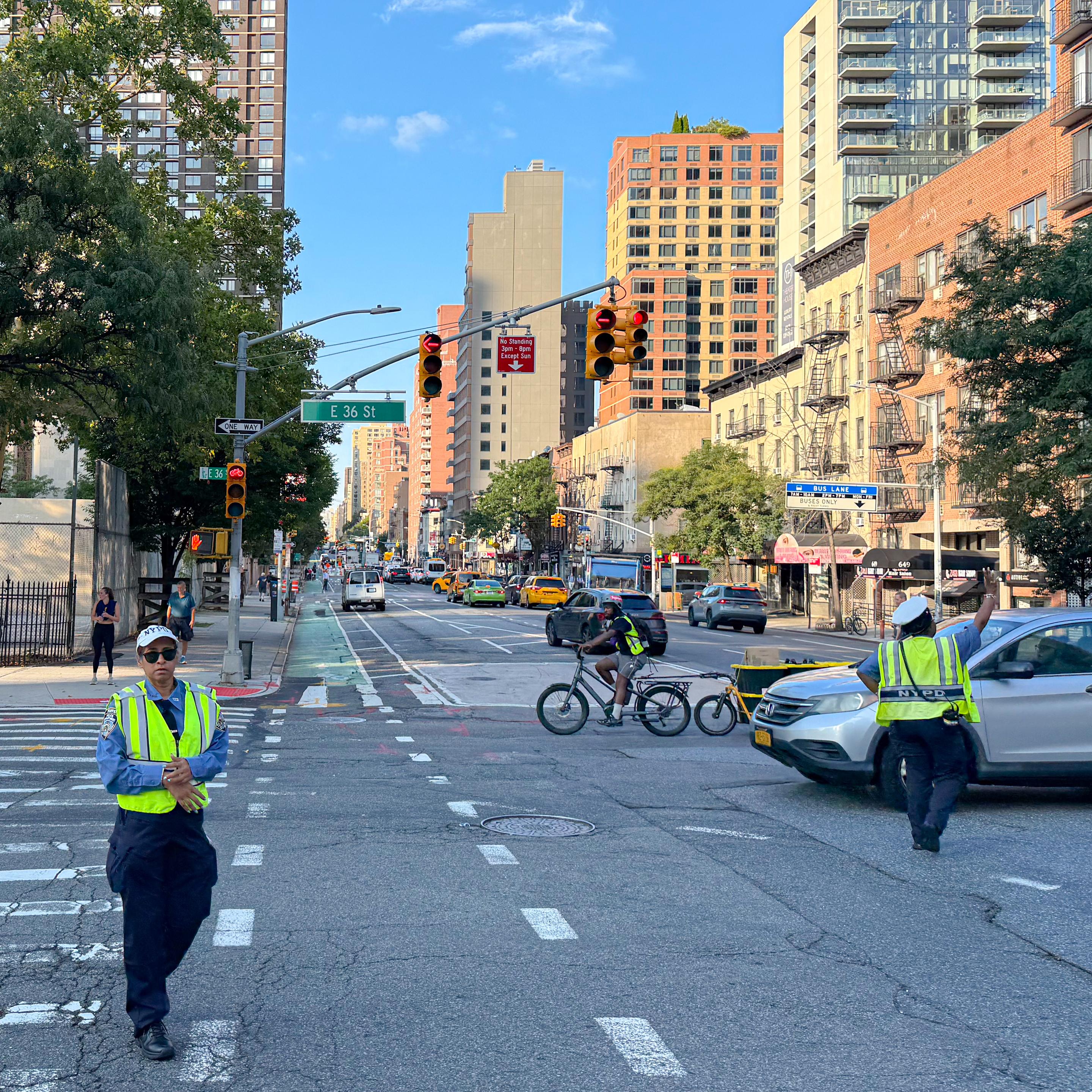 A busy urban street scene featuring traffic lights, a cyclist, and two traffic officers in reflective vests directing vehicles. Various buildings line the street, along with visible taxis and green trees. The sky is clear with a few clouds.