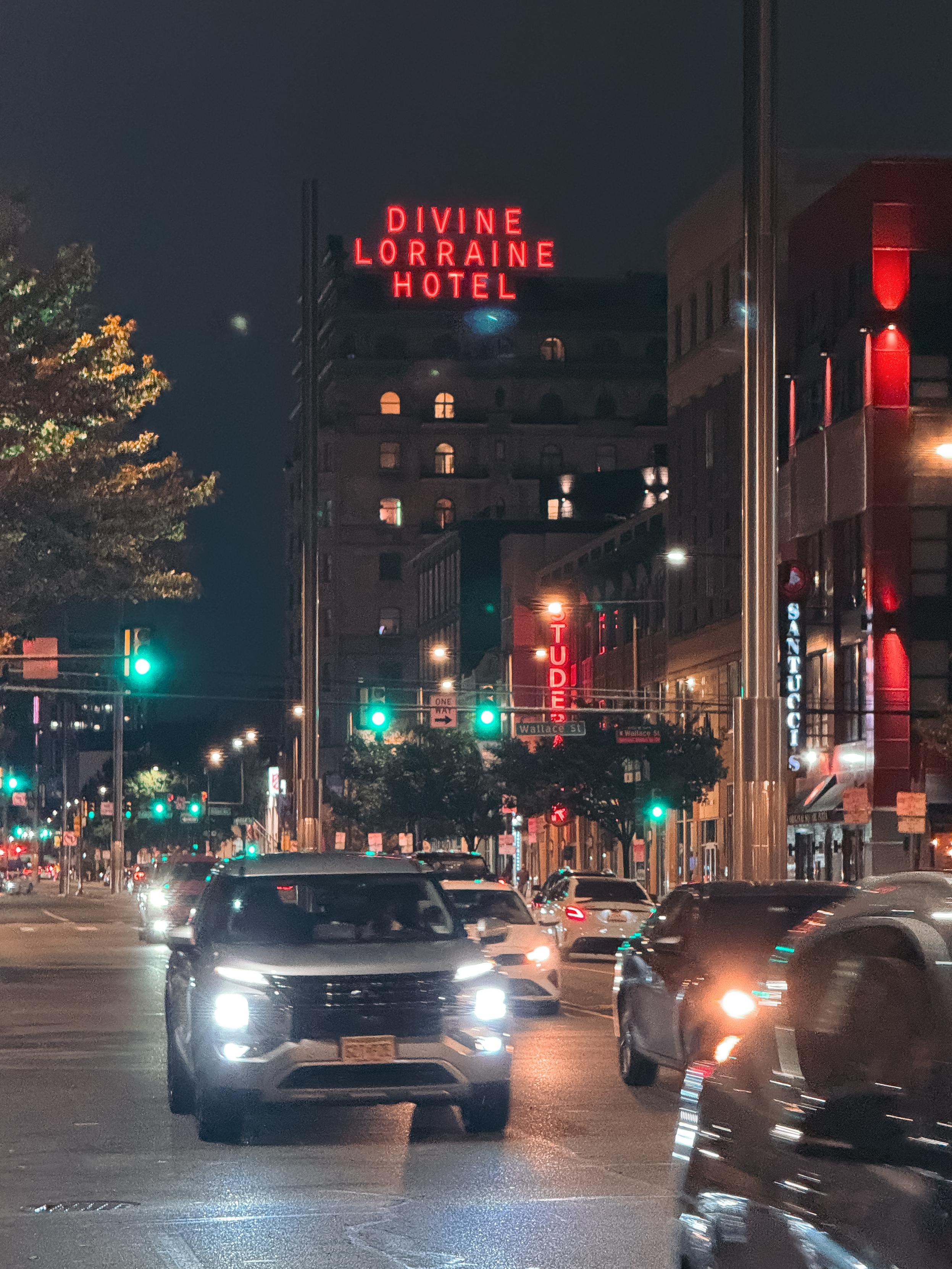 Nighttime city scene featuring illuminated traffic signals and the "Divine Lorraine Hotel" sign in red against a dark sky. Multiple vehicles are seen on the street, with shops and buildings lining the boulevard.