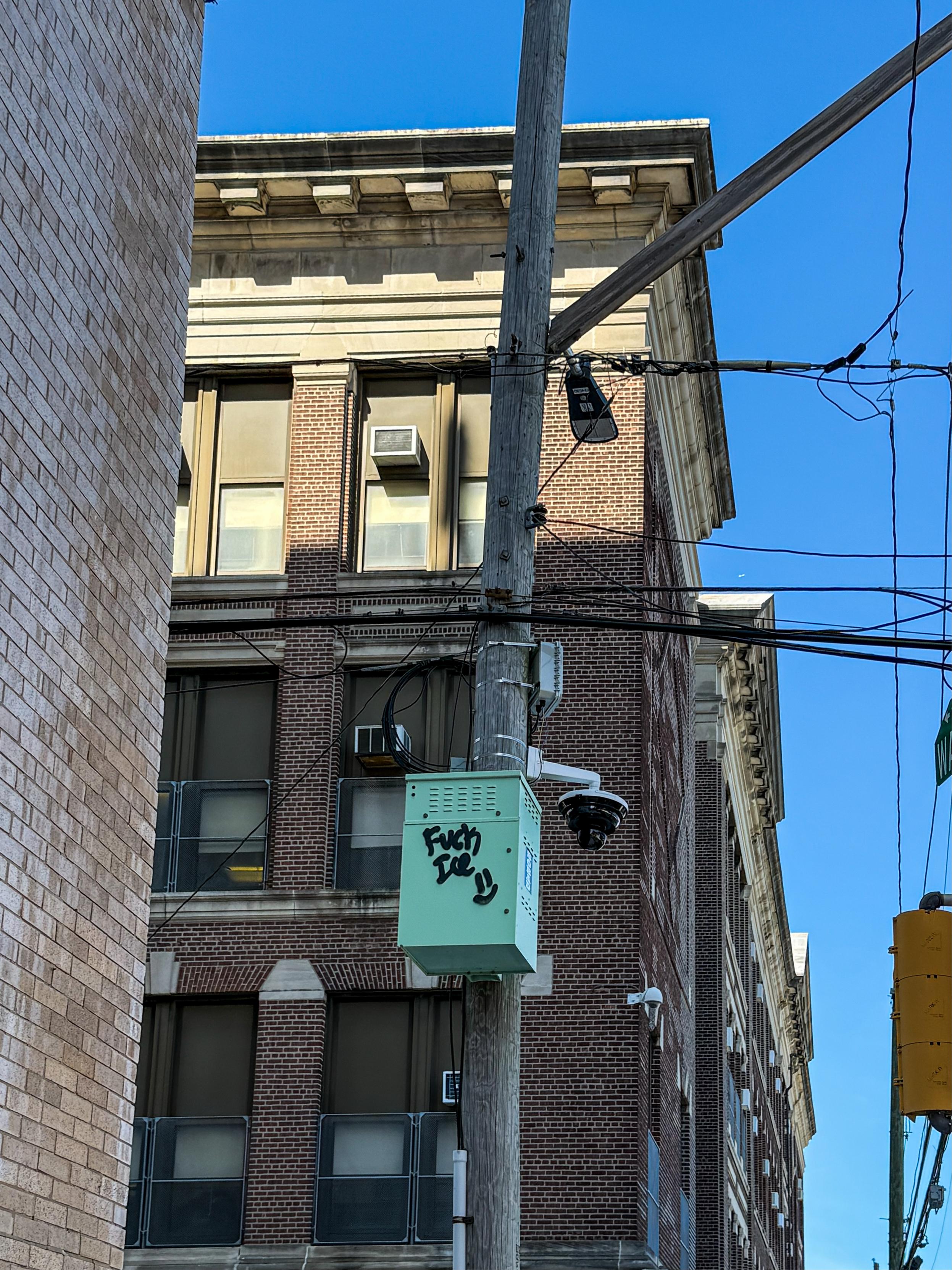 A graffiti-tagged green electrical box with the text "Fuck ICE" is mounted on a utility pole, accompanied by a surveillance camera. The background features a brick building and a clear blue sky.