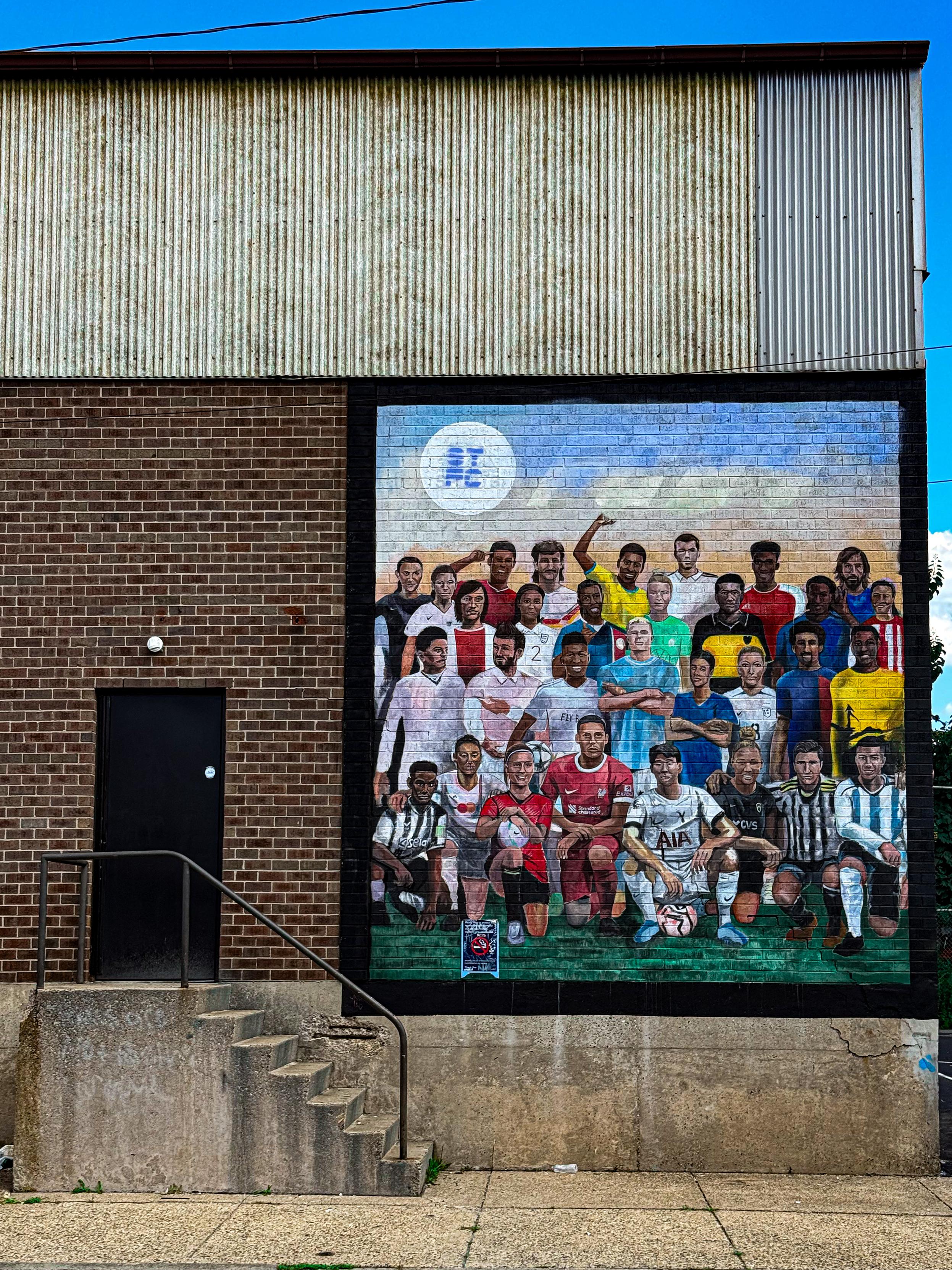 A large mural on a brick wall features a group of diverse soccer players in various kits, representing different teams and nationalities. The mural includes a mix of posed players, with some standing and others kneeling, set against a blue sky background.