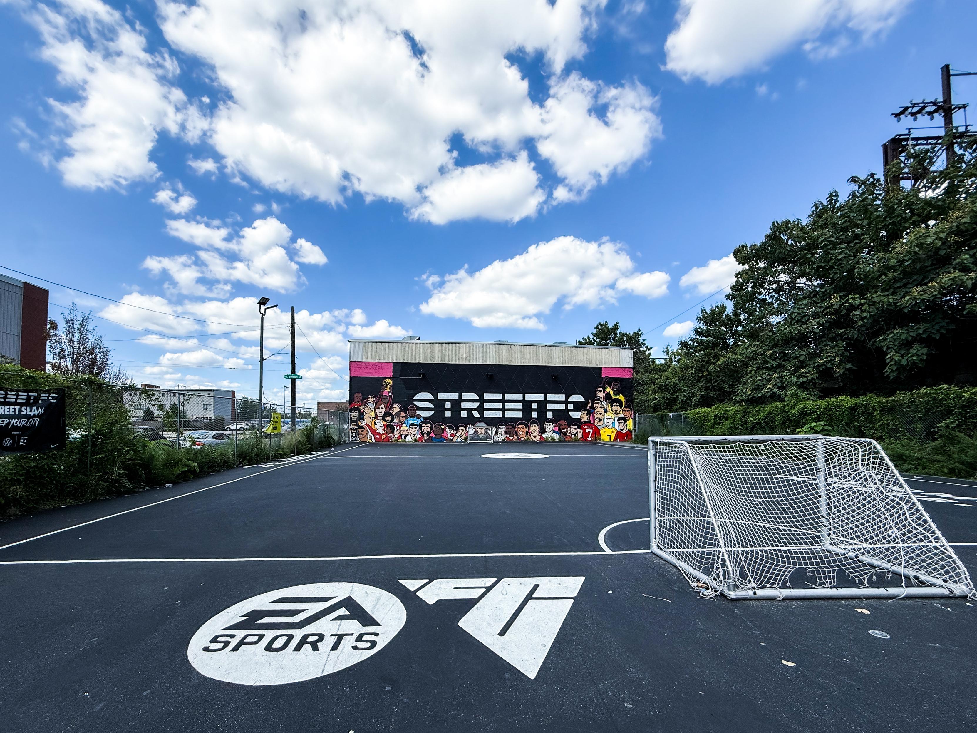 An outdoor basketball court features a large mural on the back wall, depicting colorful characters. A small goalpost is positioned on the court, with an "EA Sports" logo visible on the ground. The sky is filled with fluffy clouds.