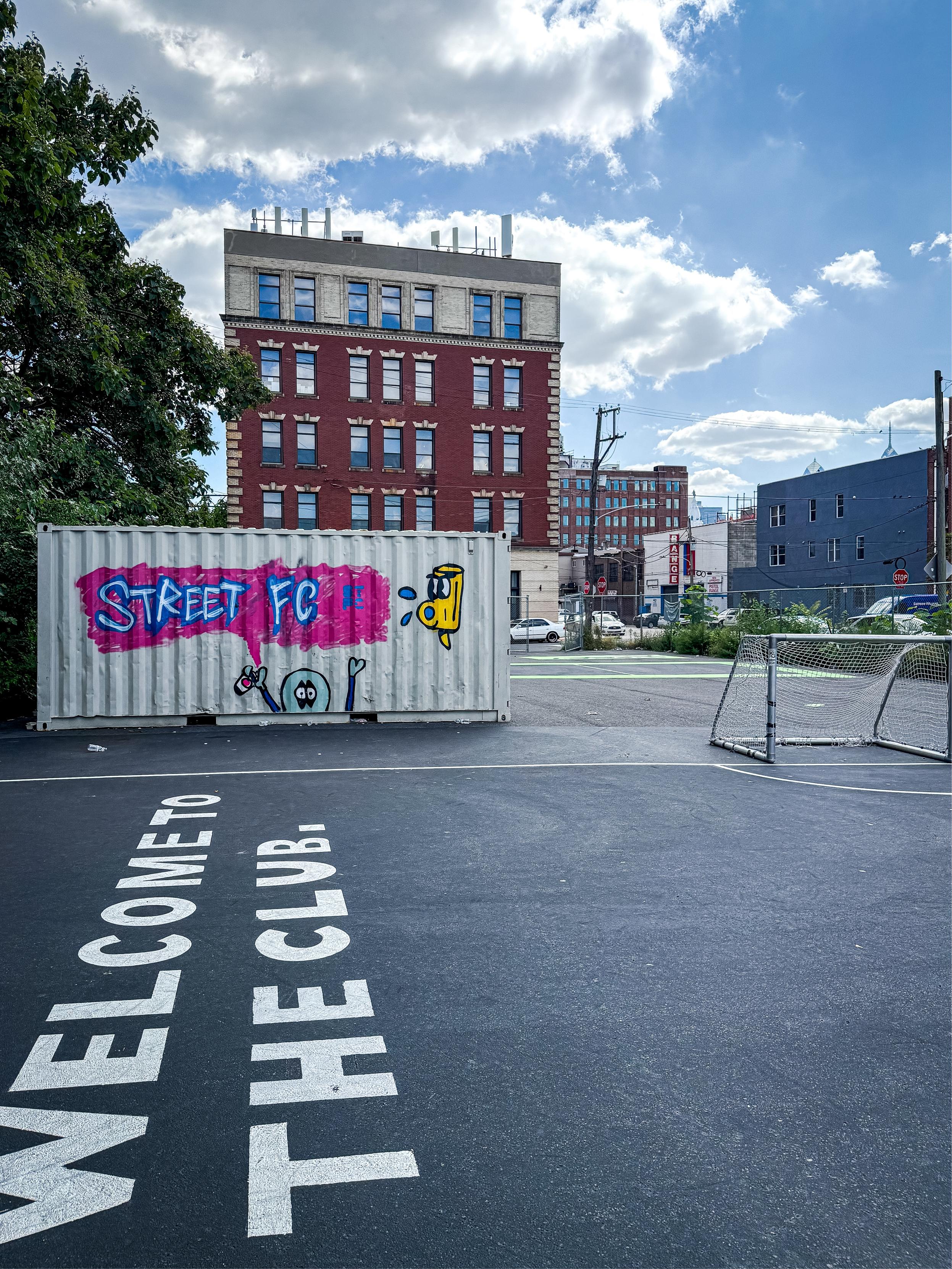 A shipping container featuring colorful graffiti labeled "STREET FC" sits in an open area with a partially paved surface. The text "WELCOME TO THE CLUB" is painted on the ground in large white letters, with a backdrop of a red-brick building.