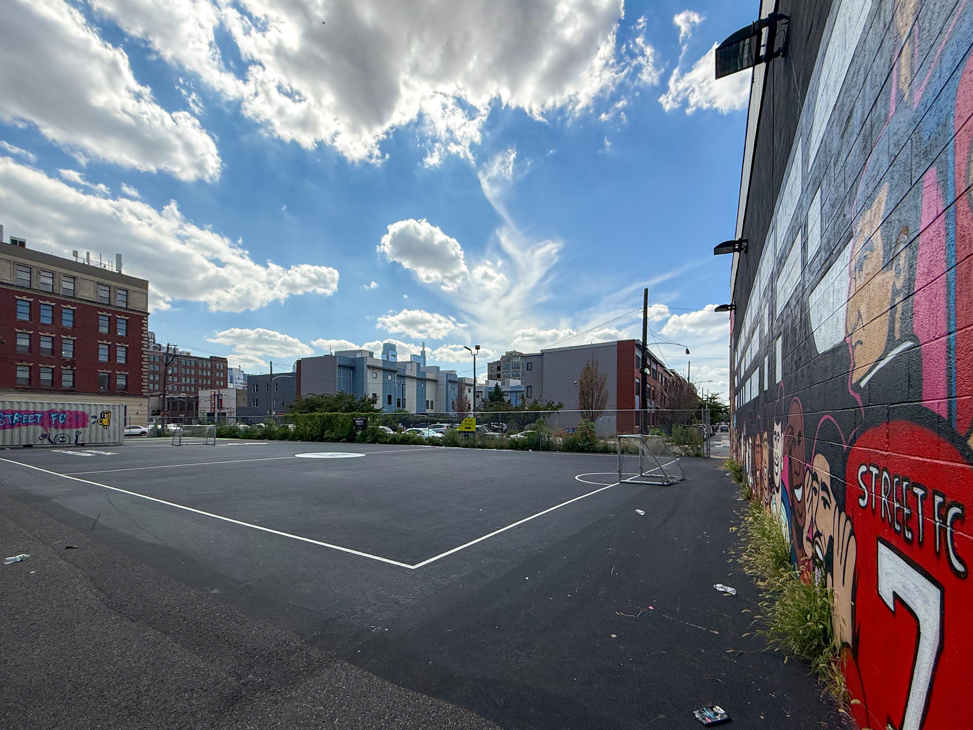 An outdoor soccer court with a graffiti-covered wall in the background. The scene features cloudy skies and a mix of urban buildings. There are goals on the court and some greenery along the edges.