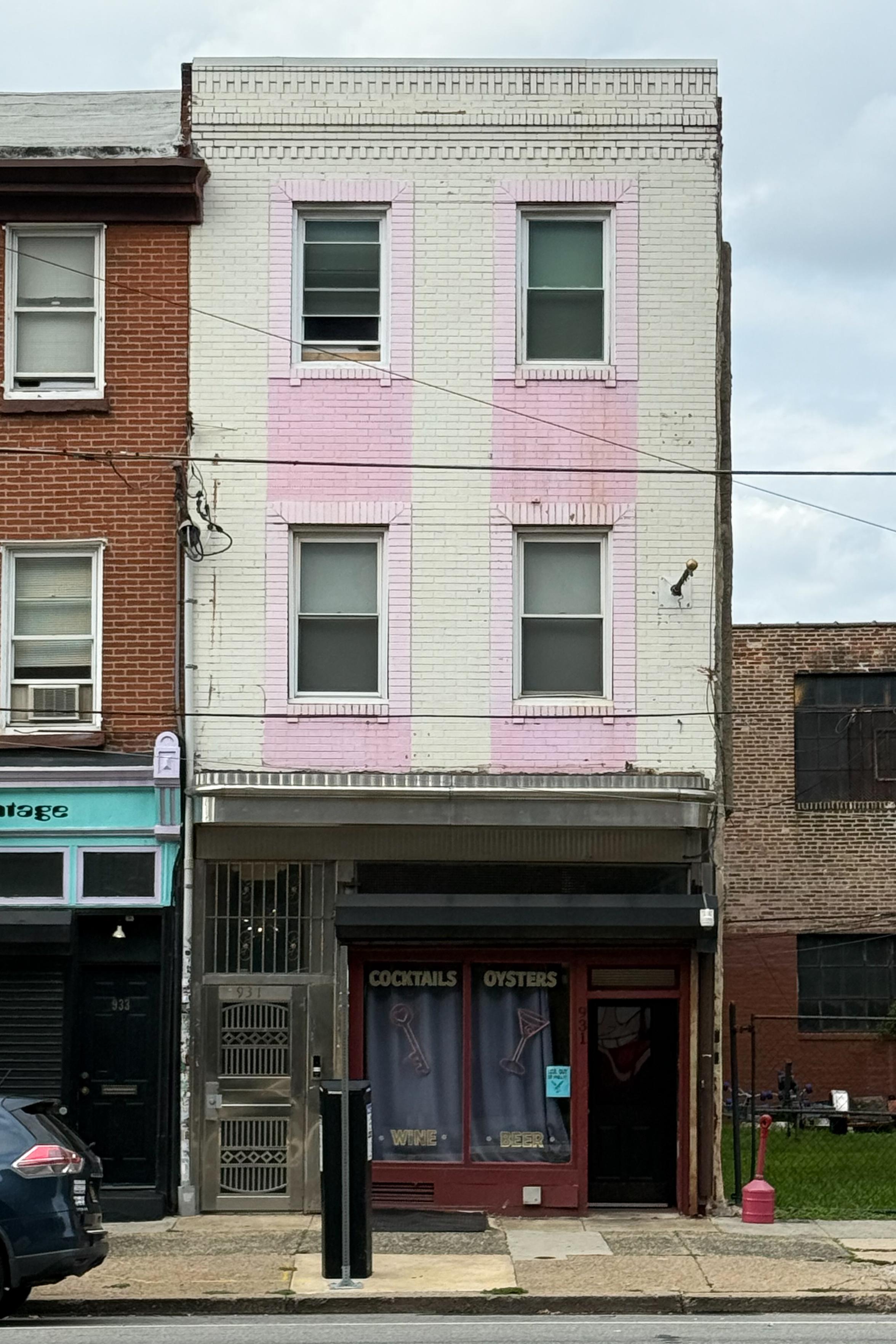 A three-story building with a mix of brick and painted white and pink exterior. The ground floor features a storefront with the words "Cocktails Oysters" on window signage. Nearby, there's another business with an aqua-colored awning.