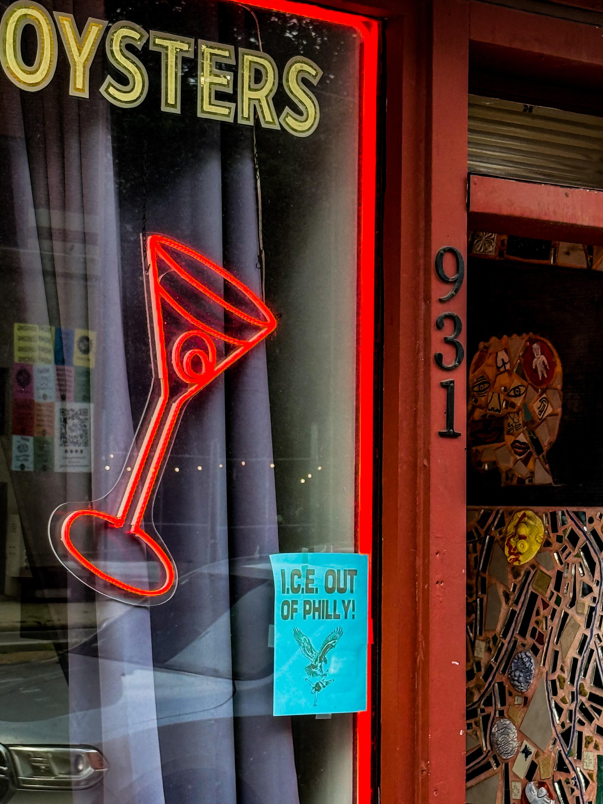 A storefront featuring a neon sign that reads "OYSTERS" and a red martini glass. A blue flyer stating "I.C.E. OUT OF PHILLY!" is visible in the window. The door frame has a mosaic design.