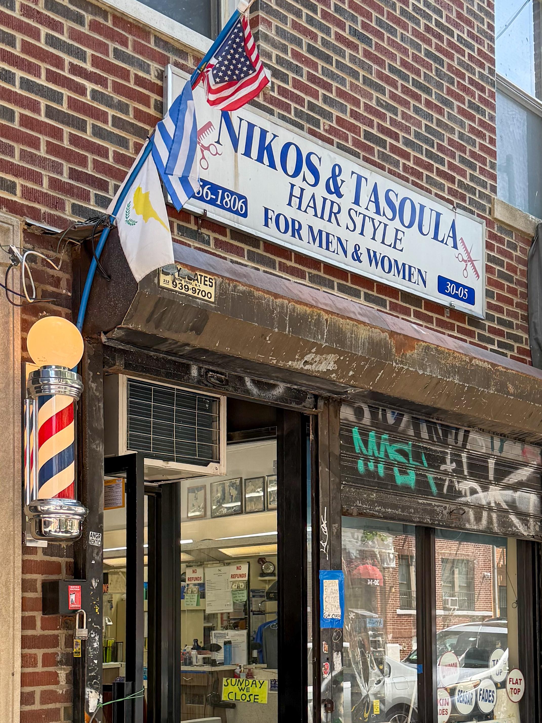 Exterior of a barbershop named "Nikos & Tasoula Hair Style" featuring a sign indicating services for men and women. Flags of the United States, Greece, and Cyprus are displayed. A traditional barber pole is visible next to the entrance.