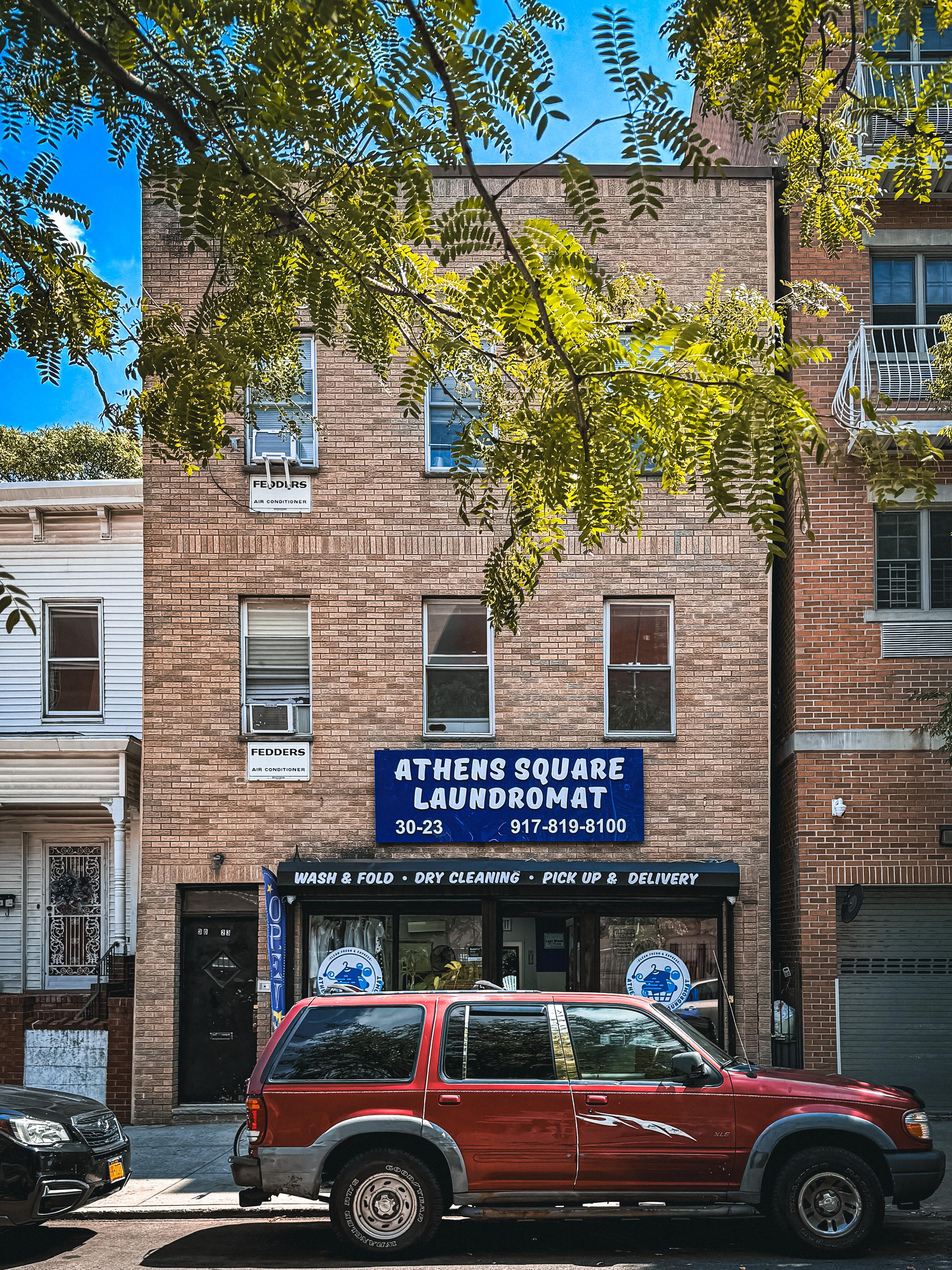 A brick building featuring a laundromat sign that reads "Athens Square Laundromat." The storefront promotes services like wash and fold, dry cleaning, and pickup and delivery. A red SUV is parked in front, and leafy trees frame the shot.