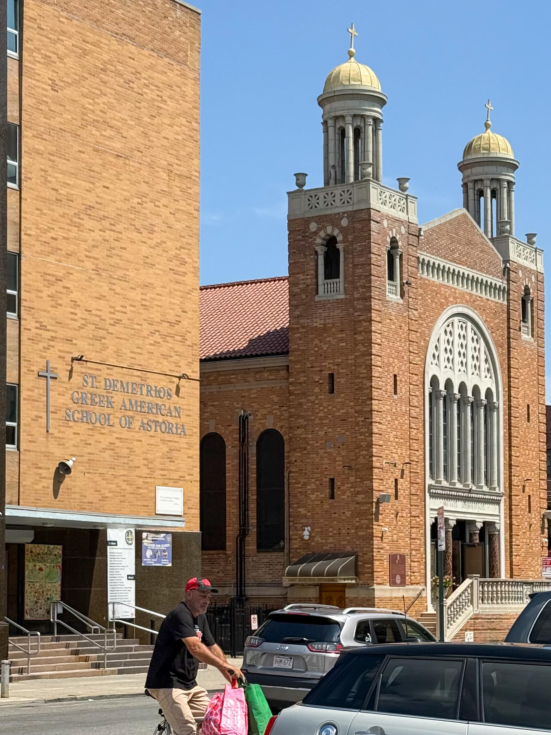 The image features St. Demetrios Greek American School of Astoria beside a distinctive Greek Orthodox church building with domed towers of the same name. A man on a bicycle carrying bags is passing in front of parked cars under a clear blue sky.
