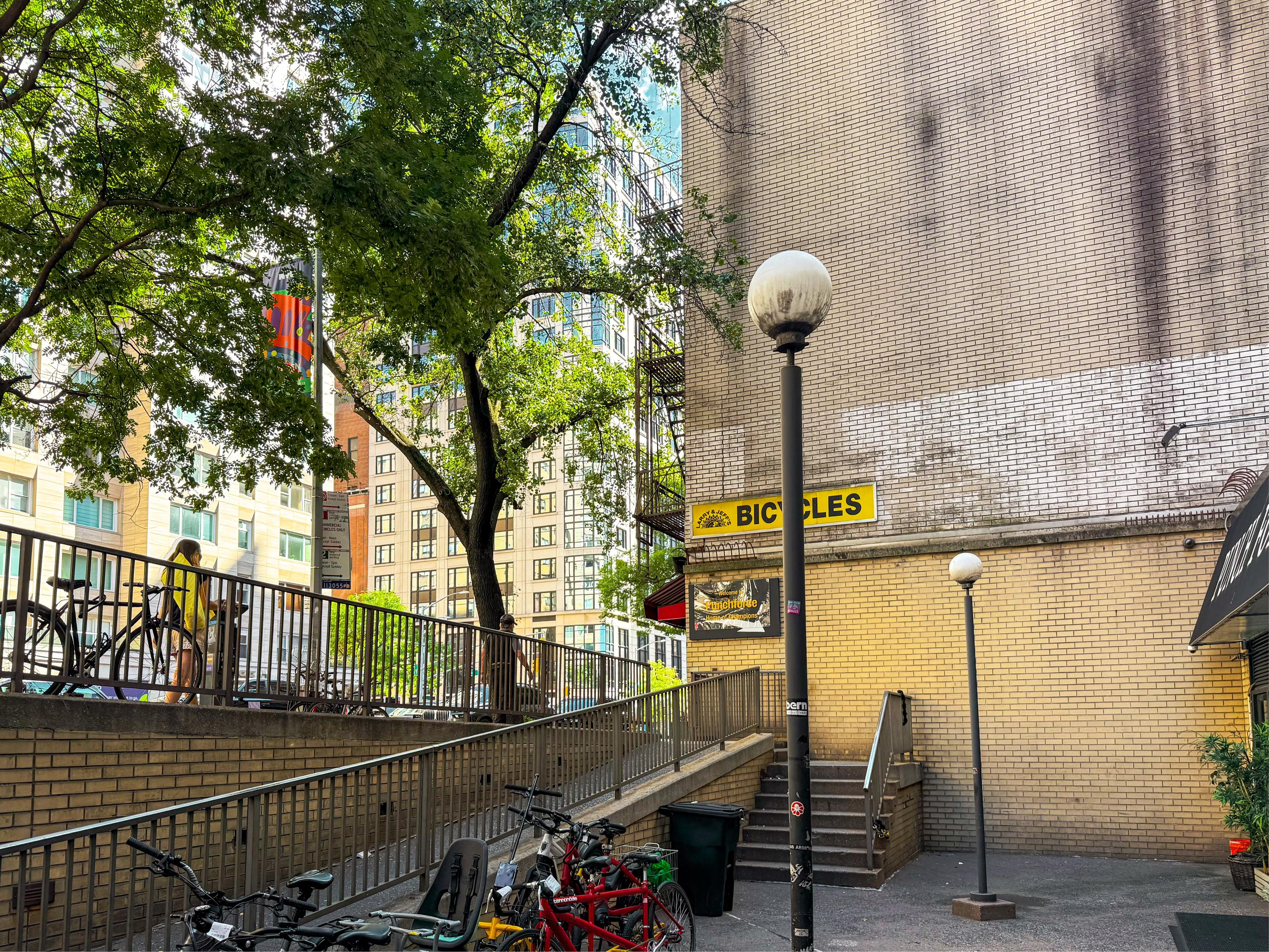 A city scene featuring a stairway leading to a building with a sign for bicycles. There are trees providing shade and several bicycles parked along a railing. Bright sunlight illuminates the area, and modern buildings are visible in the background.
