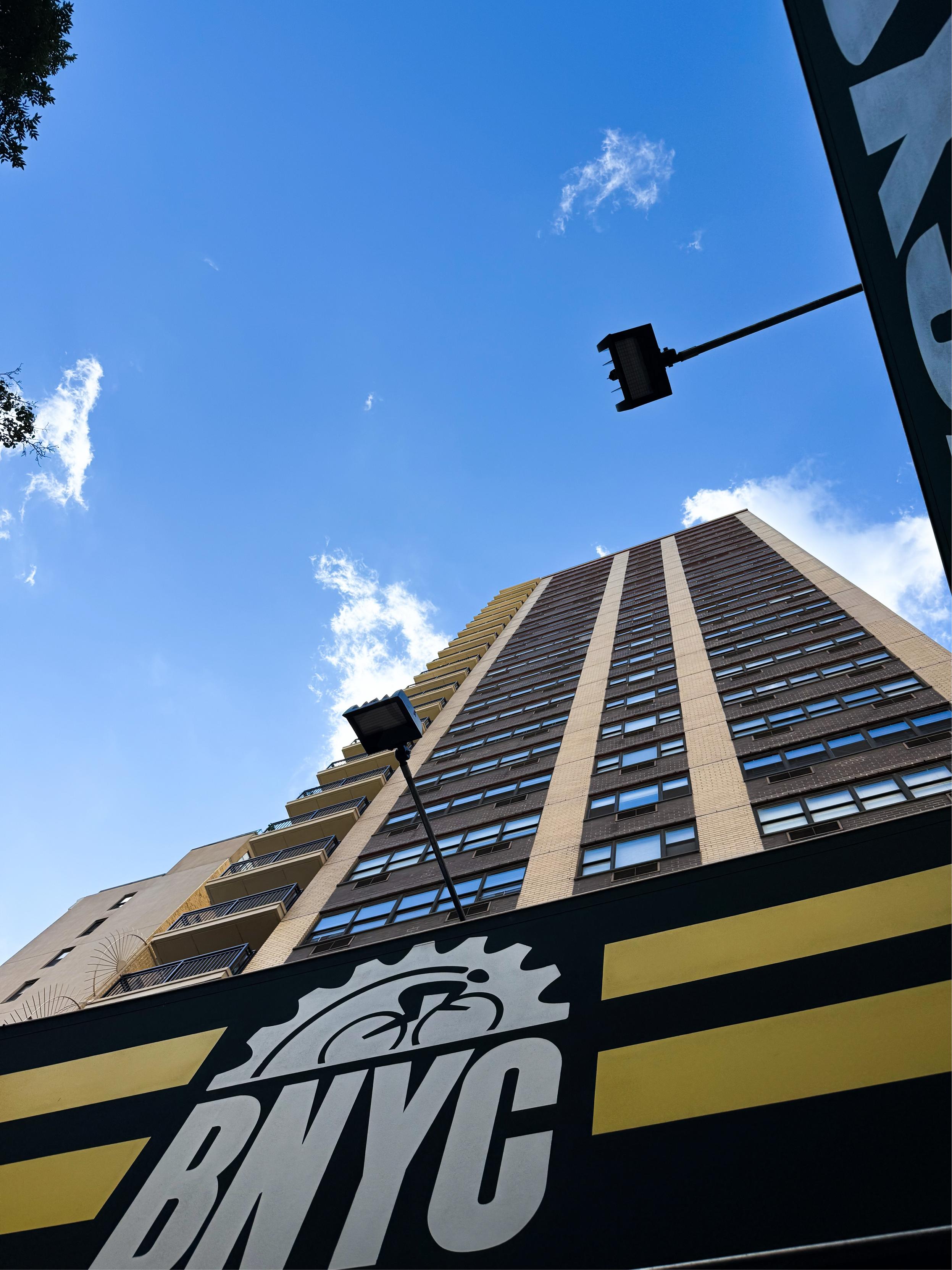 A tall building reaches into a clear blue sky, with a sign displaying "BNYC" and a bicycle graphic at the bottom. The scene is framed by greenery on one side and street lighting above.
