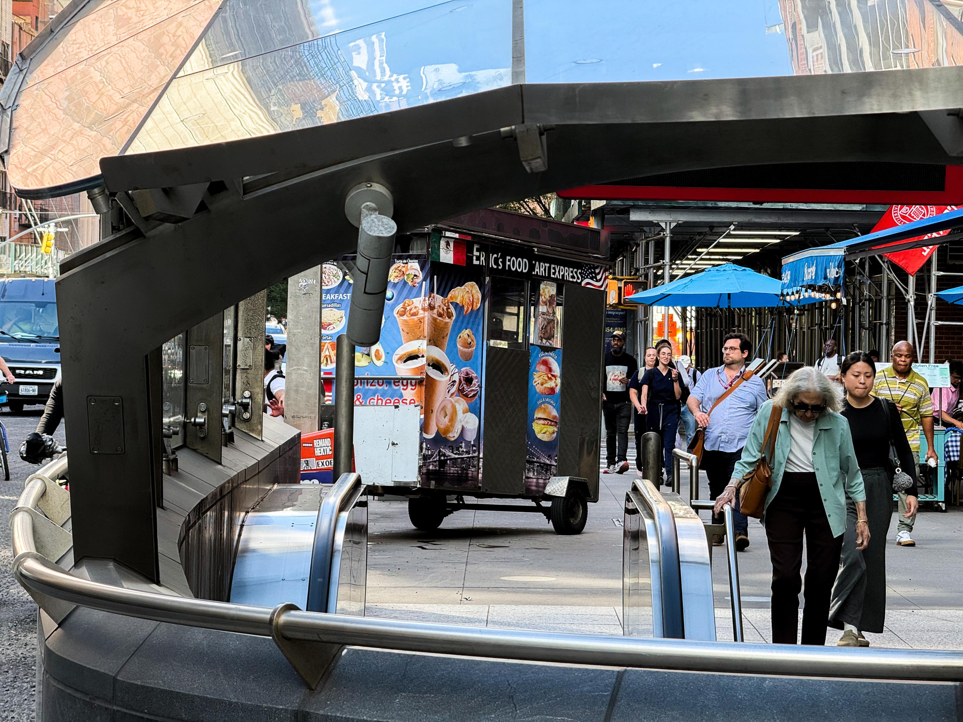 A busy street scene featuring people walking past a food cart labeled "Ice Cream Food Art Express." There are steps and a railing in the foreground leading to the 72 Street Subway Station below, with a diverse group of pedestrians and blue umbrellas and awnings.