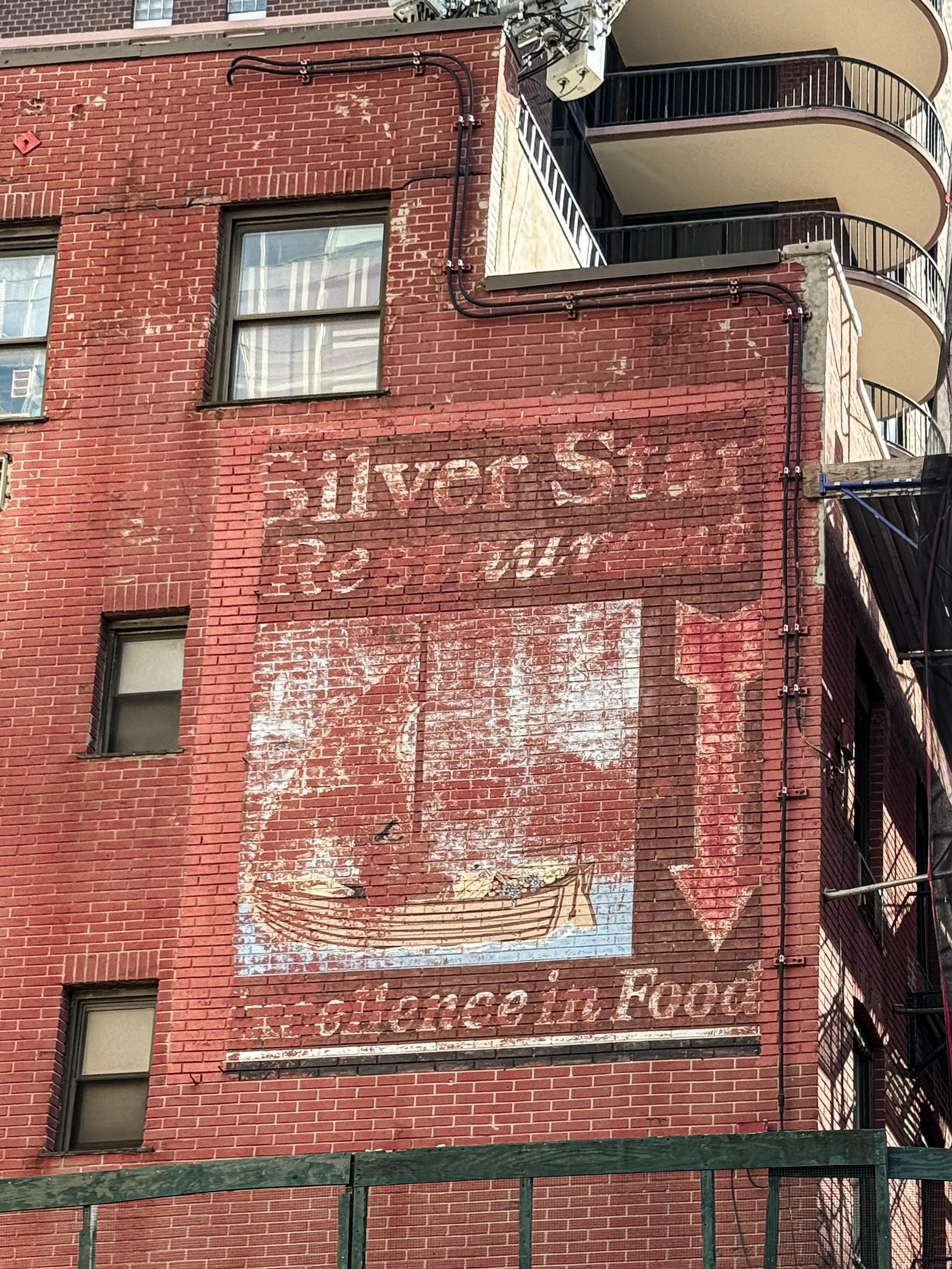 Faded mural on a brick wall featuring the text "Silver Star Restaurant" and an image of a boat, with a downward arrow and the phrase "Excellence in food."