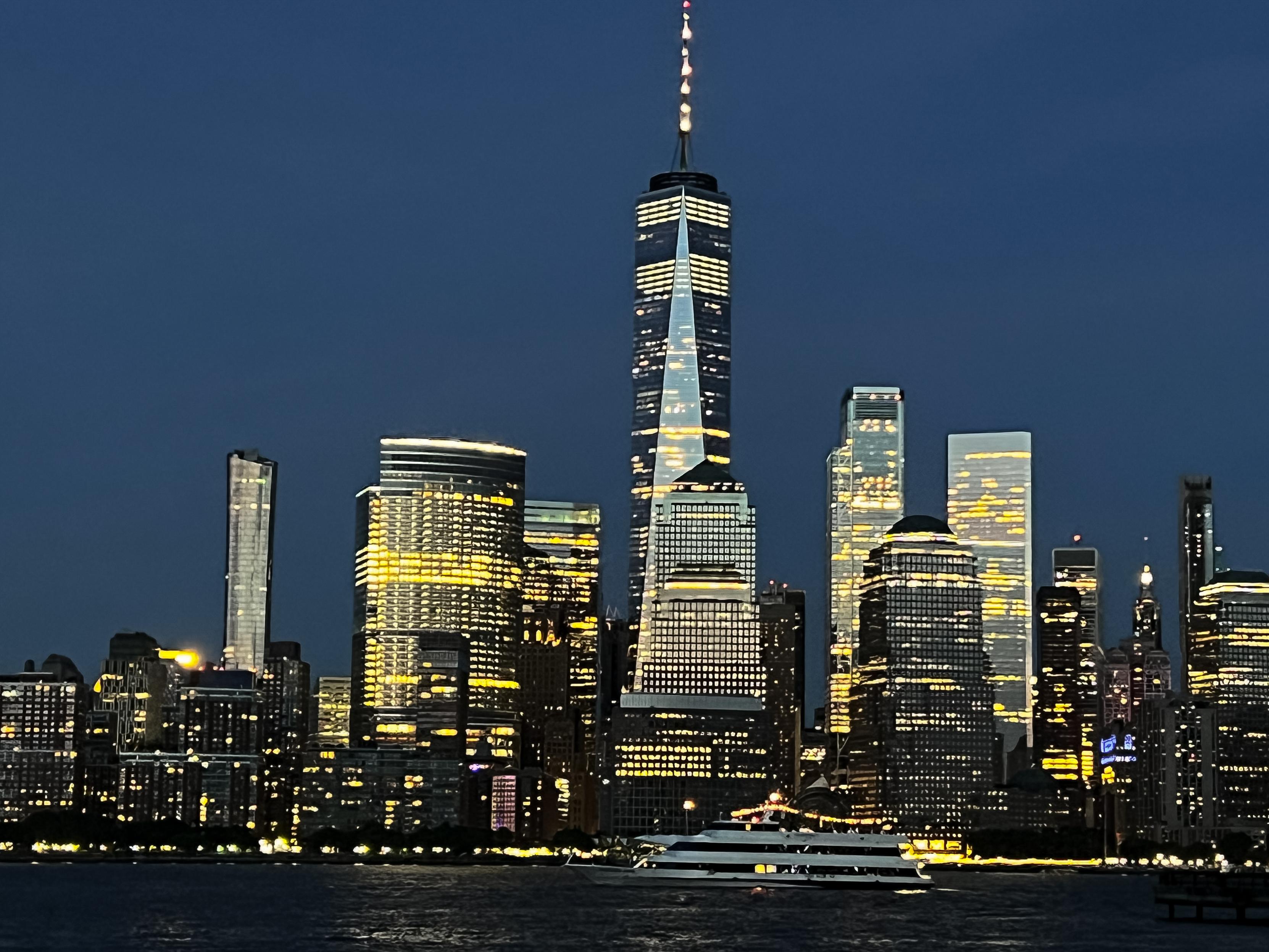 A night view of part of Manhattan’s skyline with illuminated skyscrapers, including the prominent One World Trade. A ferry is visible in the foreground, reflecting the city lights on the water. The sky is darkening, highlighting the buildings' lights.