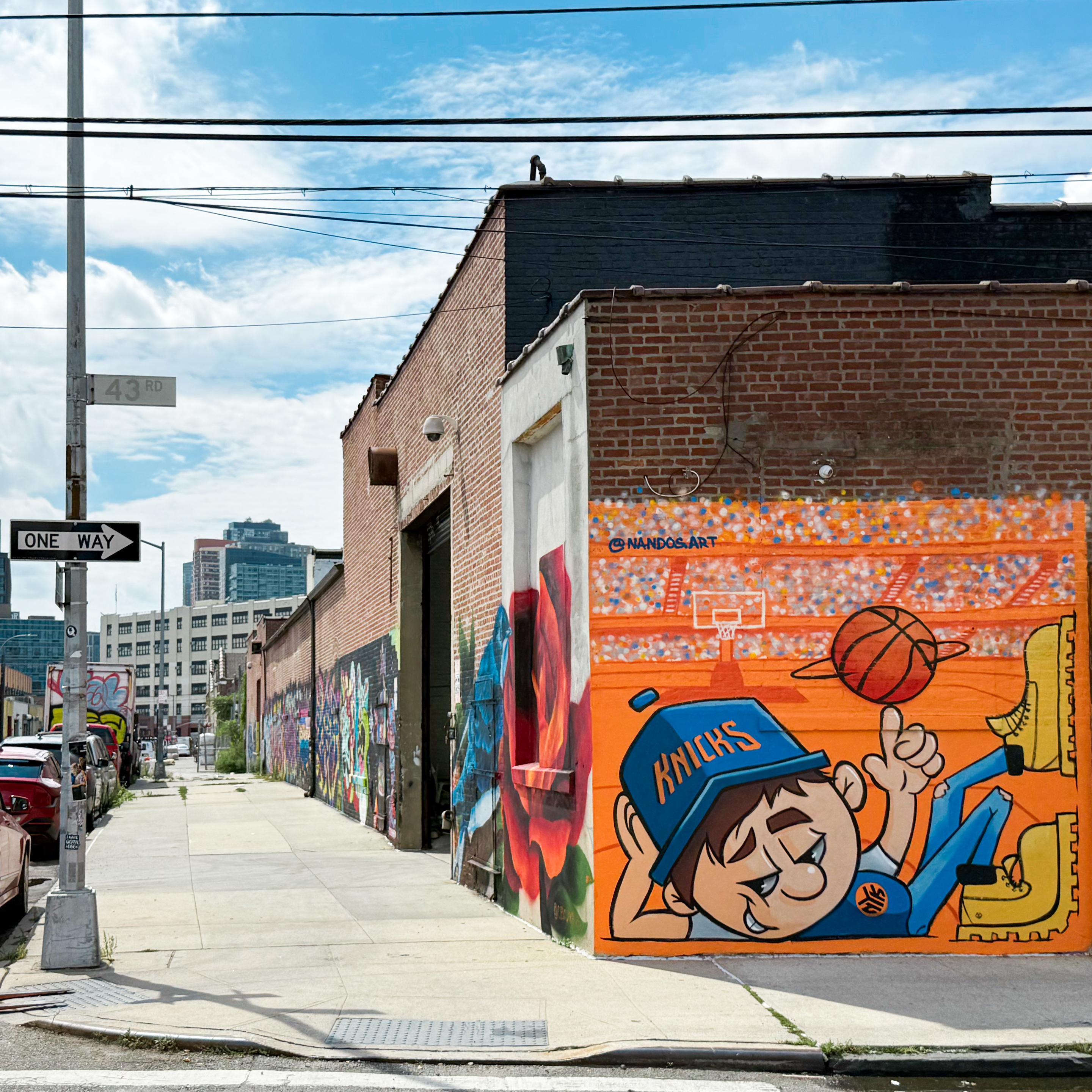 A vibrant urban scene featuring a mural of a cartoon boy in a blue NY Knicks cap, playing basketball. The mural is painted on a brick wall alongside a street that has various colorful graffiti artworks. Street signs indicate "43rd" and "One Way."