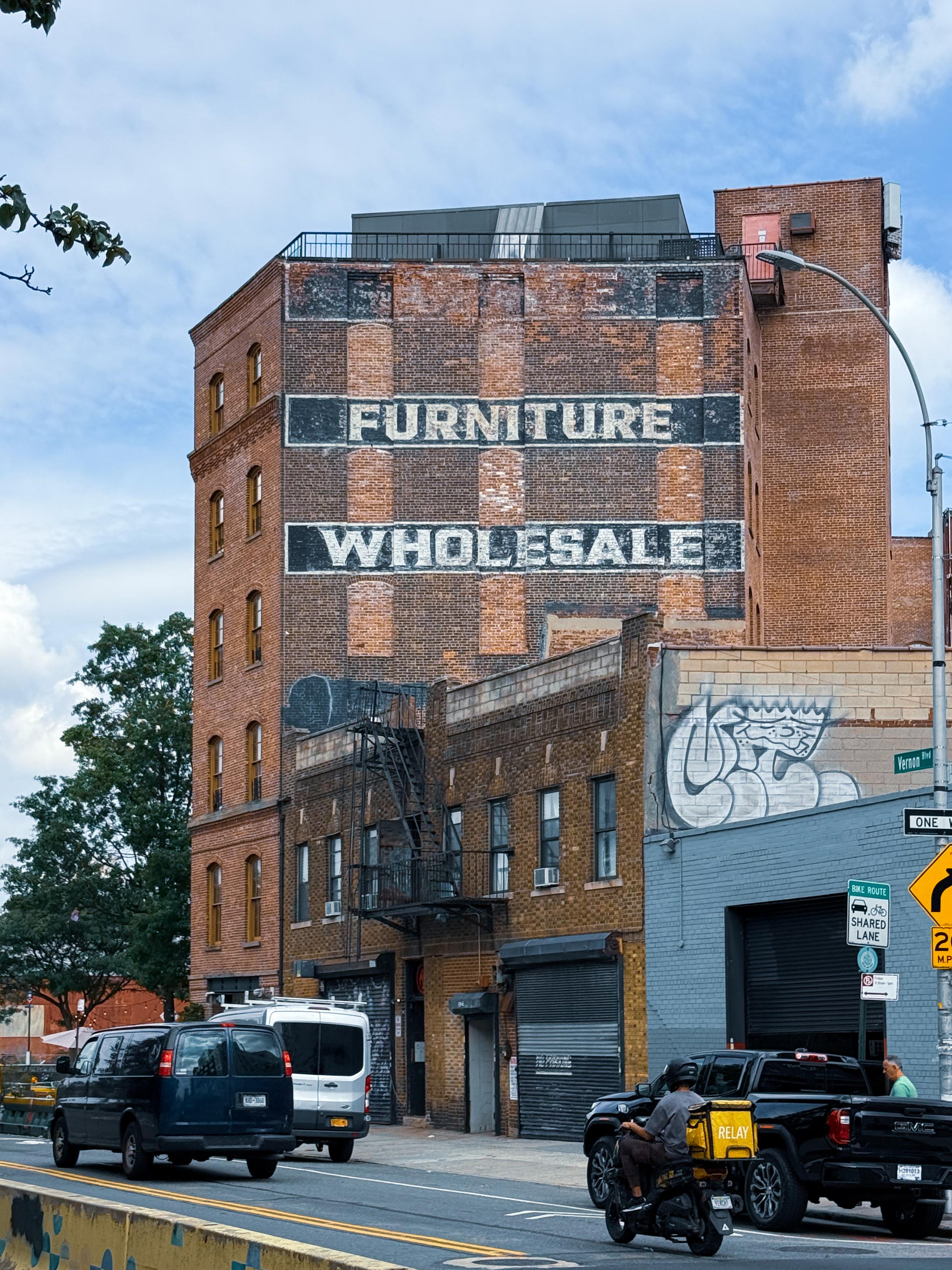 A brick building featuring faded signs that read "FURNITURE" and "WHOLESALE" on its upper sections. Nearby vehicles are visible on the street, along with graffiti on an adjacent wall.