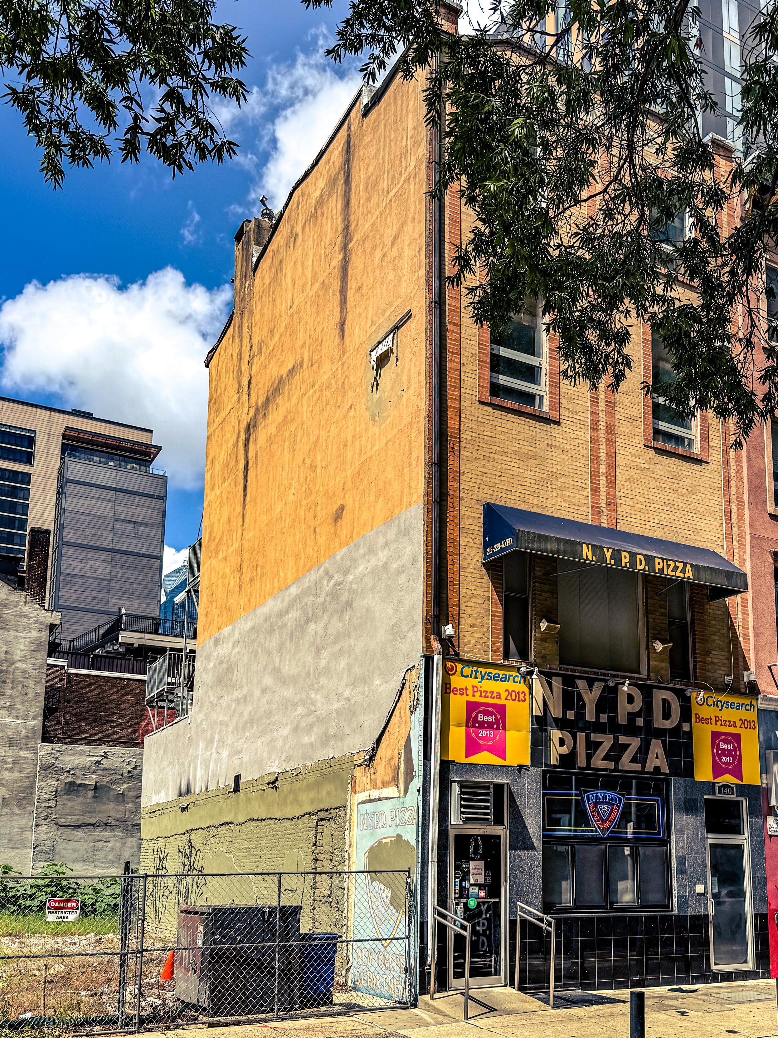 An urban scene featuring a pizza restaurant named "N.Y.P.D. Pizza." The exterior showcases a mix of orange and beige walls, with a partially visible advertisement for "Best Pizza 2013." There are nearby buildings and a fence in front of an empty lot next door.