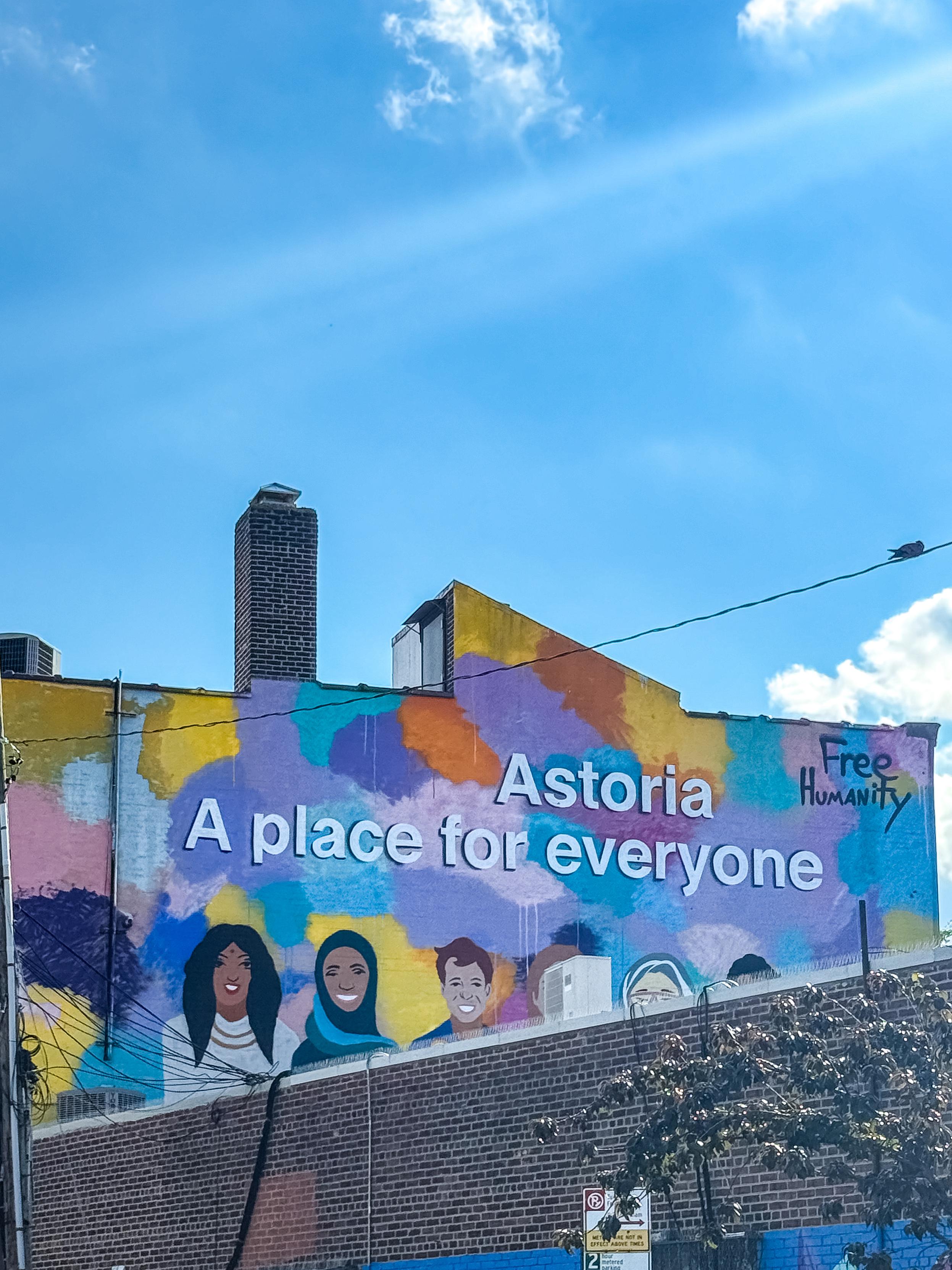 A colorful mural featuring diverse illustrations of people with the text "Astoria A place for everyone" and "Free Humanity." The background includes vibrant, abstract paint splashes against a clear blue sky.