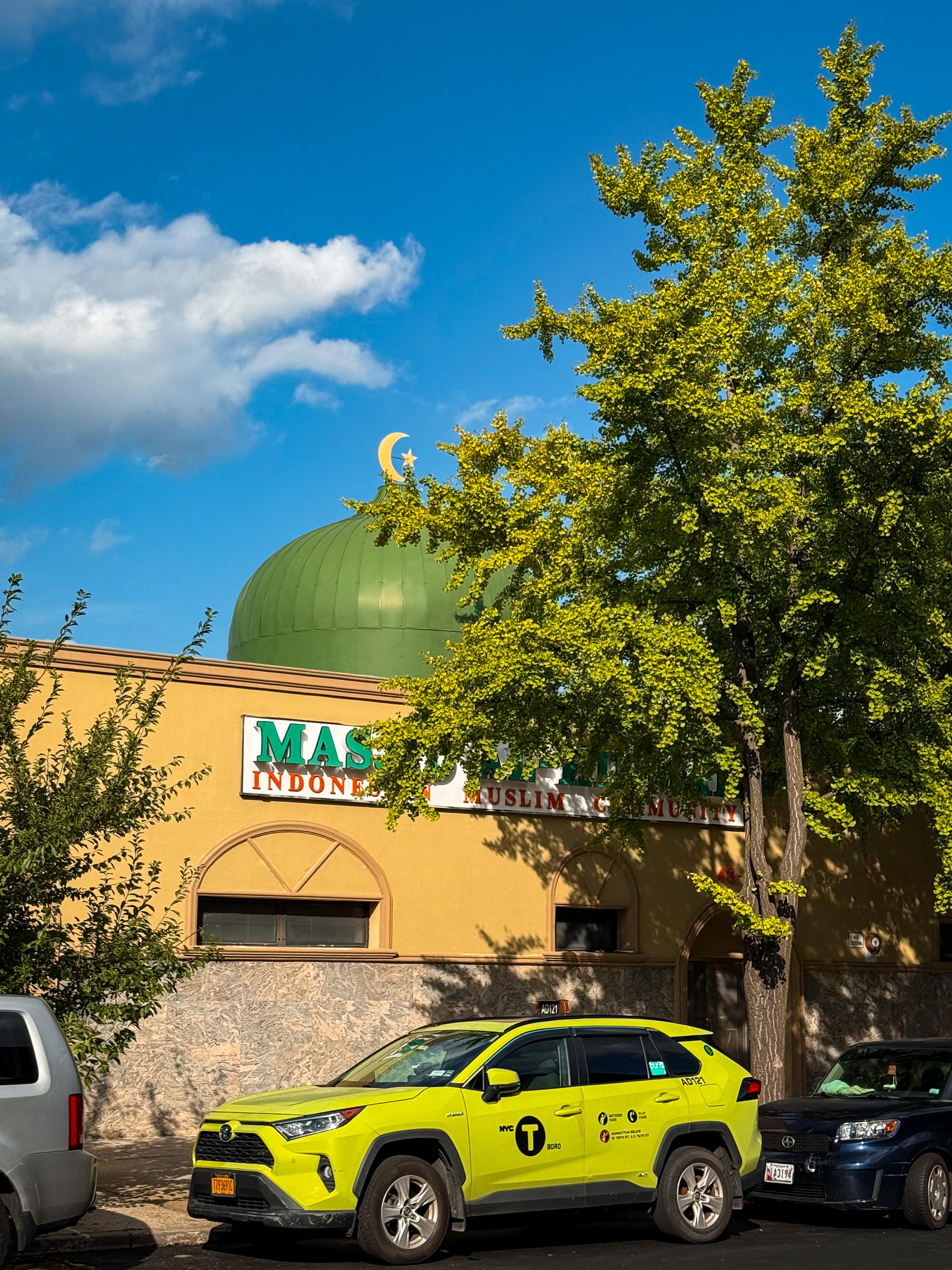 A green dome with a crescent moon and star is visible atop a building, which features a sign indicating it is a mosque for the Indonesian Muslim community. A bright lime green taxi is parked in front, surrounded by trees and a blue sky.