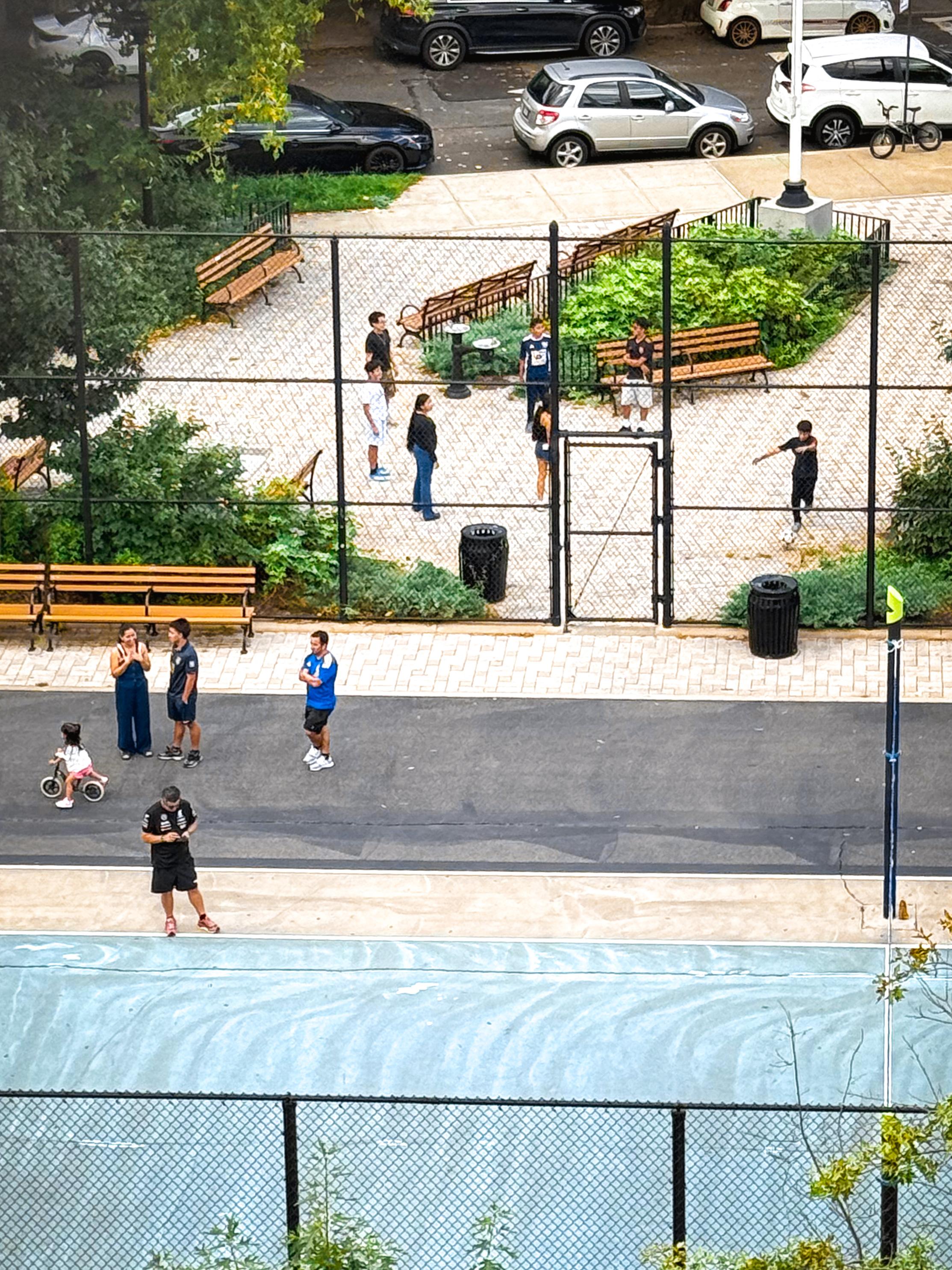 An outdoor park scene featuring several people engaged in various activities. There are benches and greenery in the background, and some individuals are playing or interacting near a fenced area. A child on a tricycle is visible, with cars parked along the edge of the park.