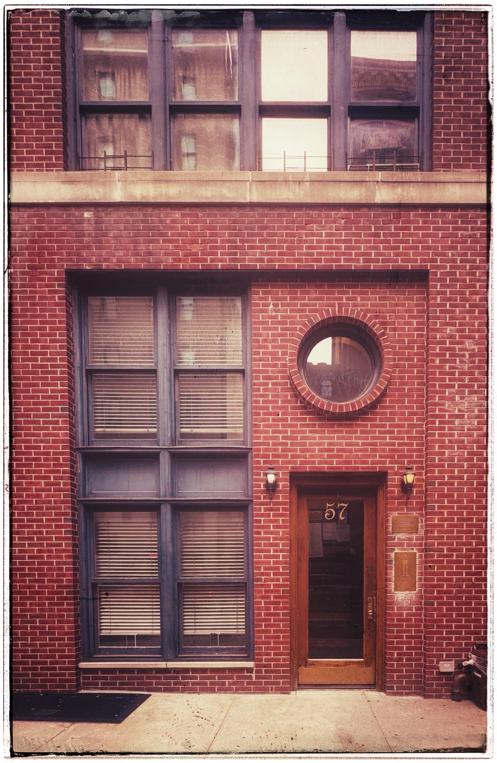 A closely cropped color photo of the front of a red brick brownstone. There's a brown wooden framed glass door on the right side with the number 57 in large gold print. Above it is a large round window outlined in brick. Next to that are two windows coming to the same height as the door and round window. Those windows have blue painted frames. On the top of the photo there is a white cement ledge and above that another set of eight square windows in horizontal rows of four,  also with the blue painted frames.