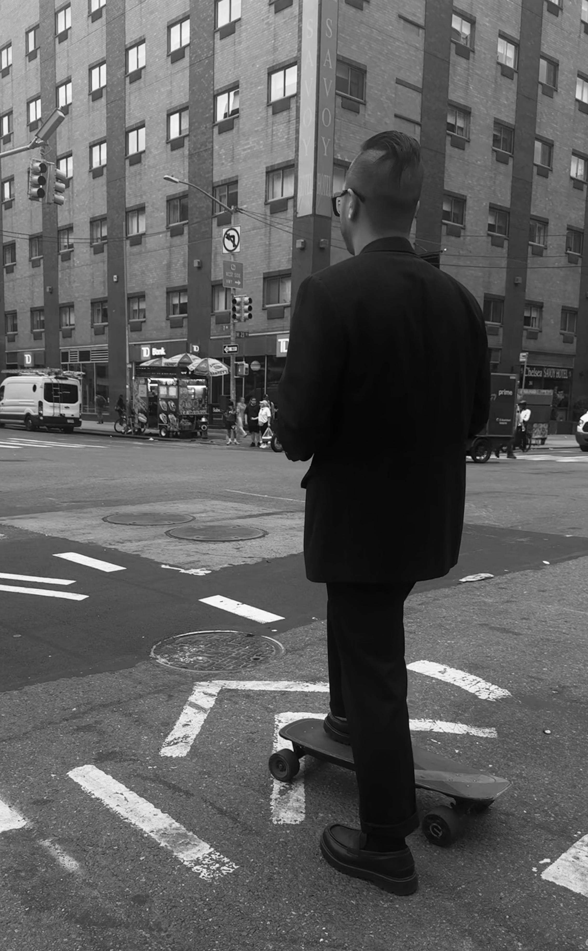 A black-and-white photo taken of a man with one foot up on a skateboard and one on the ground waiting at a red light. He is wearing cool looking black dress shoes with a full black suit, his hair is shaved except for a mohawk across the top of black hair all smoothed back and also black glasses. Behind him is a large building and the crosswalks across the street.