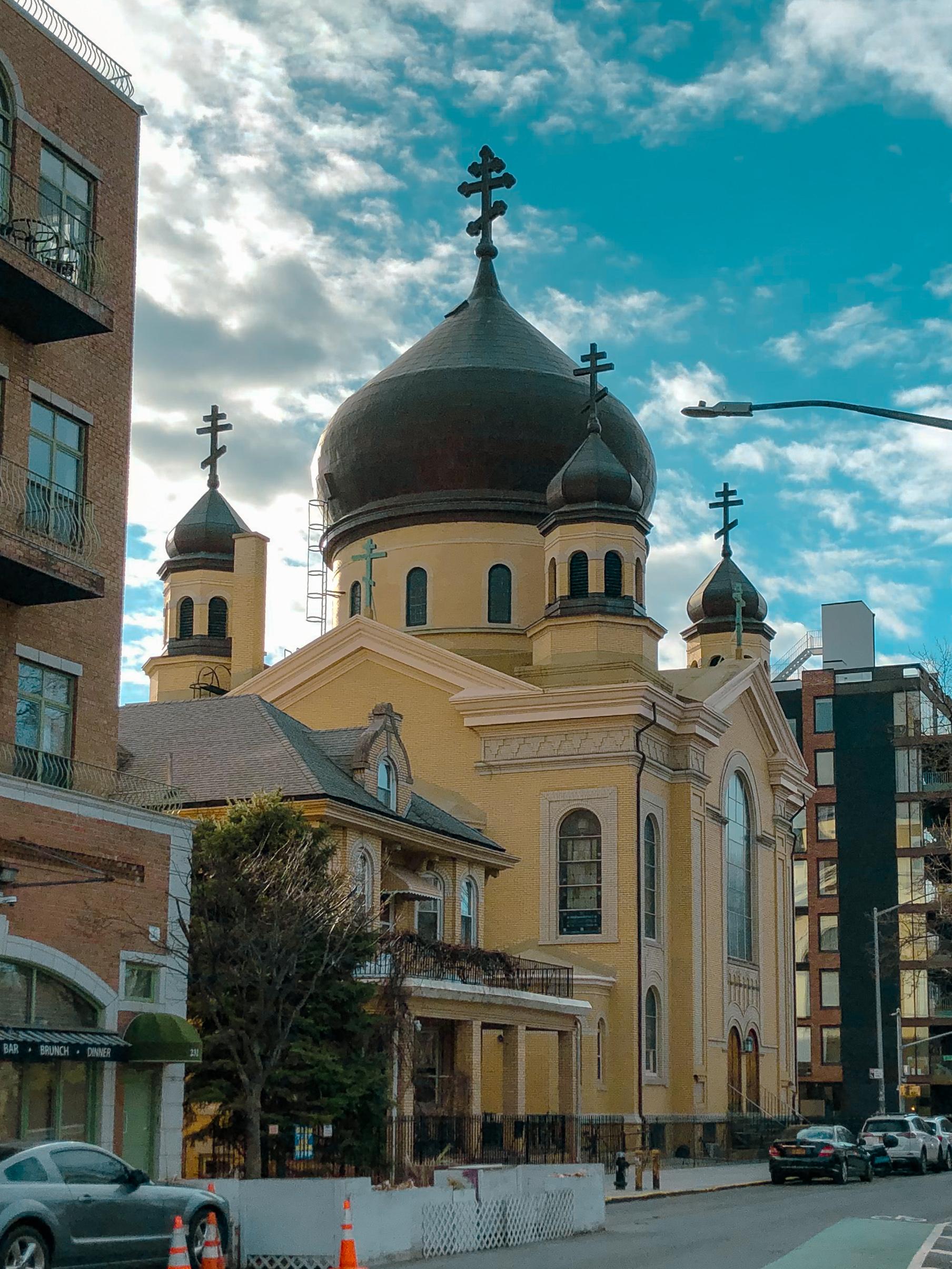 A yellow Russian Orthodox church with distinctive domes and crosses on top, surrounded by urban buildings and trees, under a partly cloudy sky.