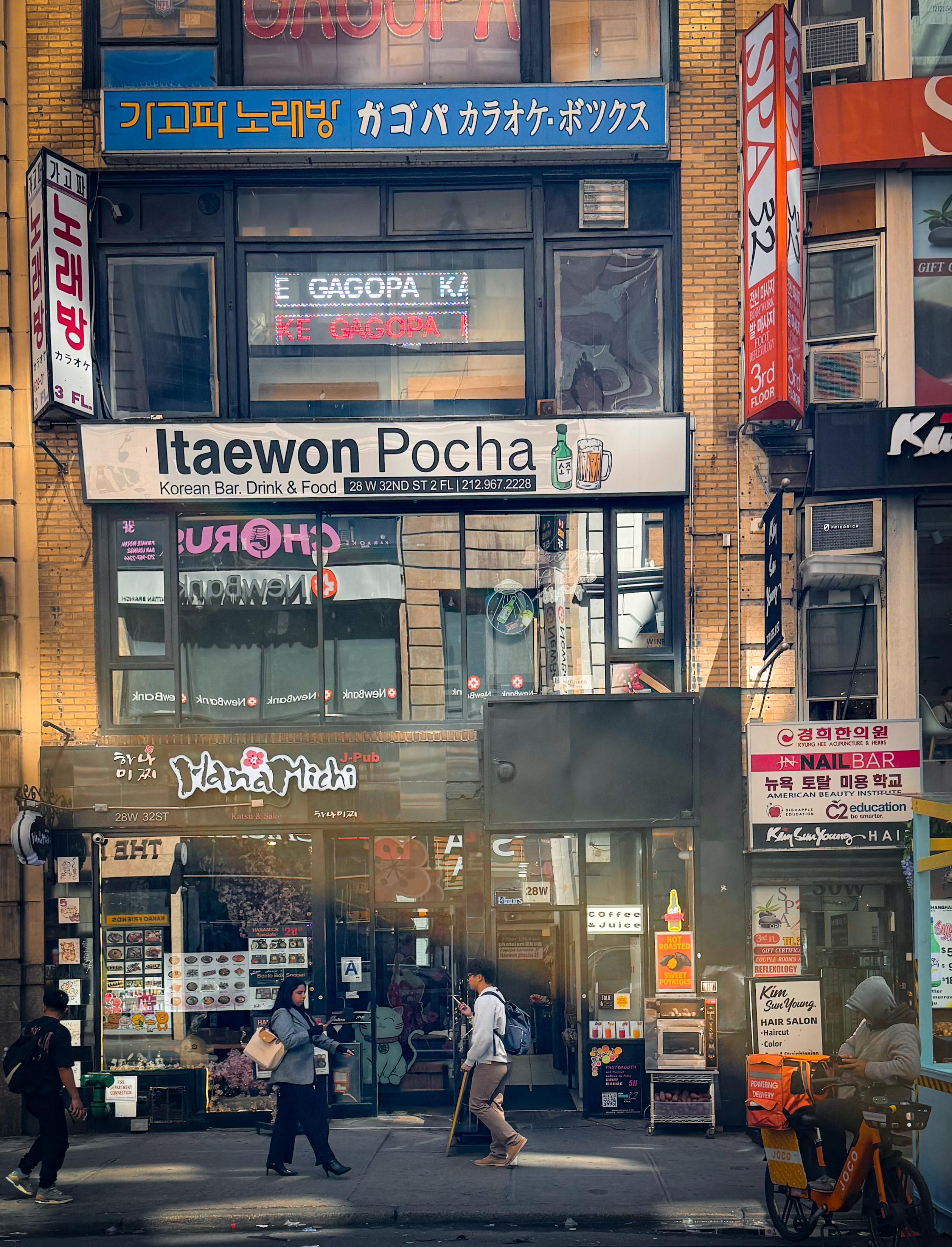 A vibrant street scene featuring multiple storefronts in an urban area. Notable businesses include "Itaewon Pocha," a Korean bar and food establishment, and "Hana Michi," a pub. The area is bustling with pedestrians.