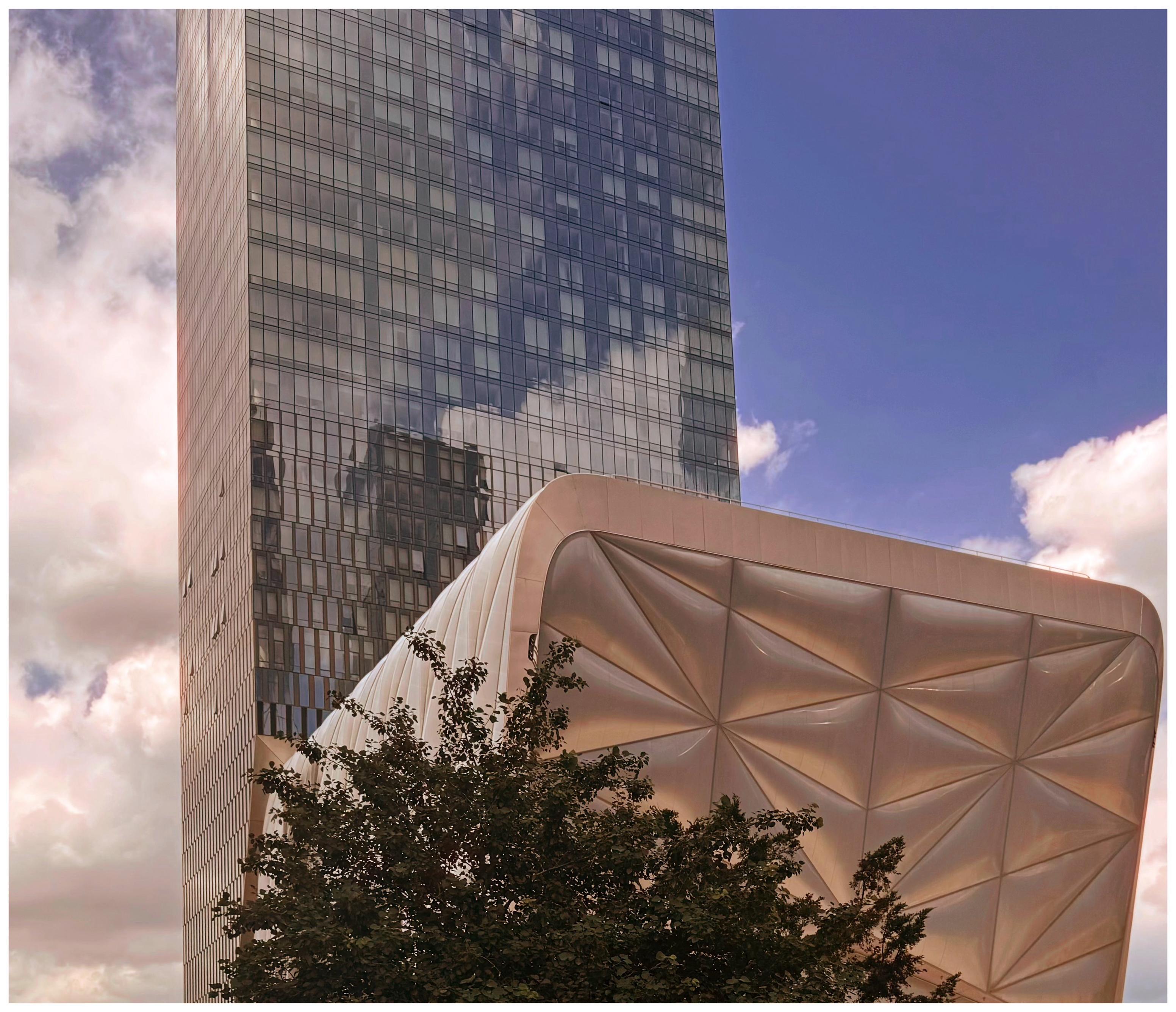 A color photo that shows the top leaves of a tree towards the bottom and above that is a square looking building that has triangles on the front that are in 3-D making it look puffy, sort of like a puffer jacket. The building is silver and with the light hitting it seems to glow a little bit. There's a large high-rise apartment building behind it with a face of small glass windows. The sky is blue on the right with more clouds down the left-hand side.