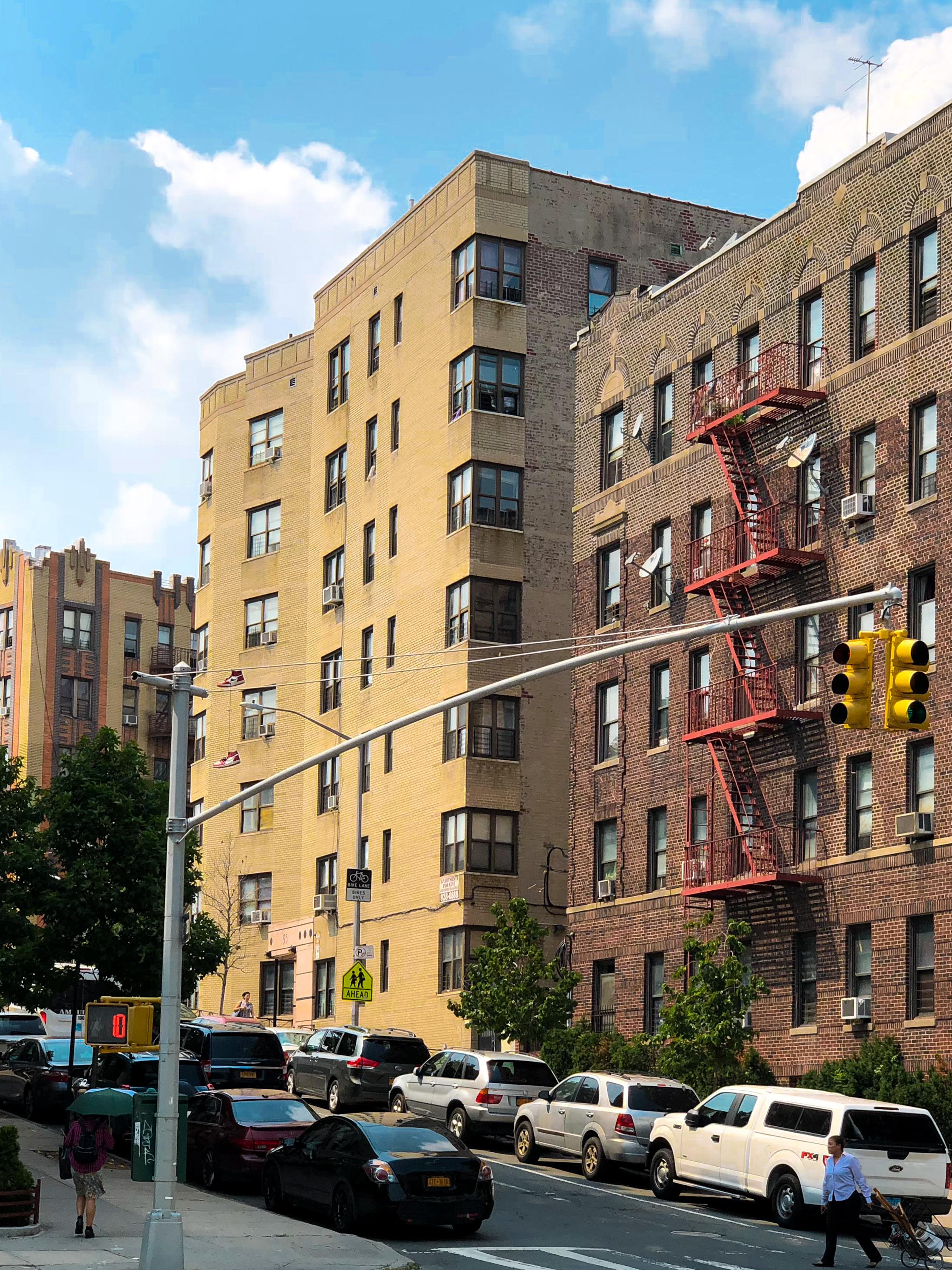 The image shows a street scene featuring two multi-story residential buildings, one yellow brick and the other brown brick with a fire escape. Parked cars line the street, and a pedestrian is walking with an umbrella. The sky is partly cloudy.