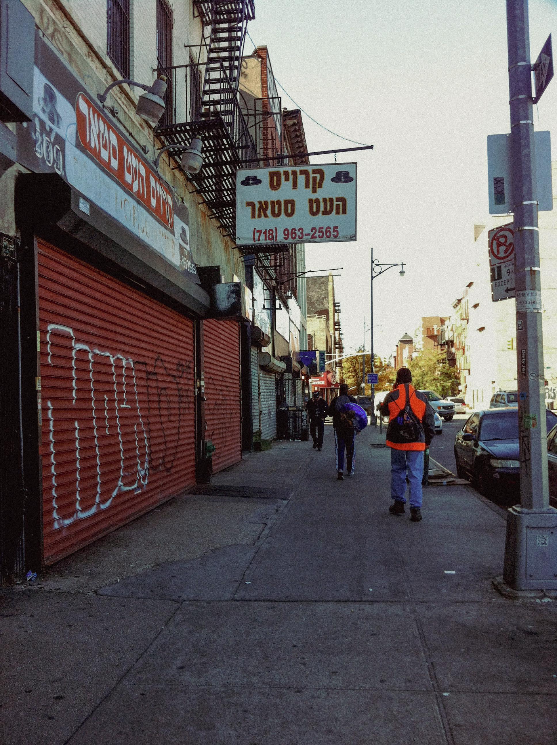 A person walking down a city sidewalk past a closed storefront with a red roll-down gate and a sign with Hebrew text.