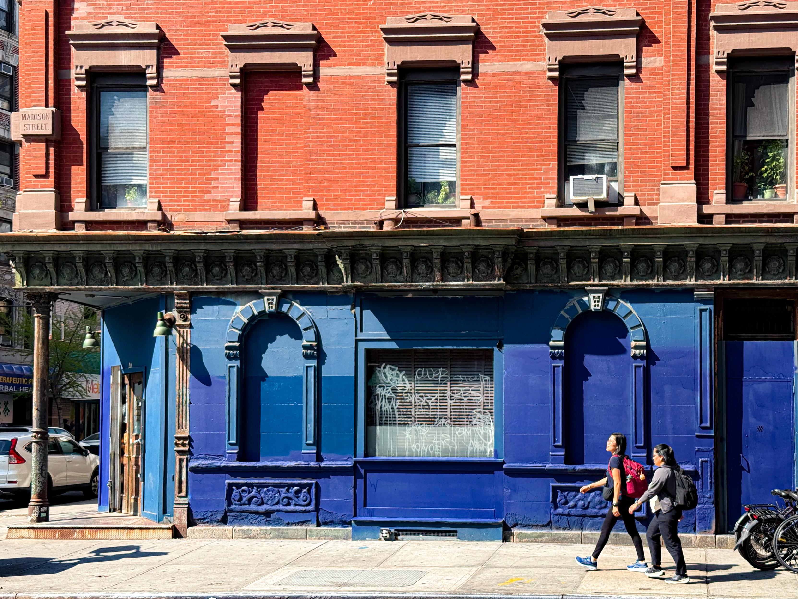 Two people walking past a vibrant blue building with a red brick upper level on a sunny day.