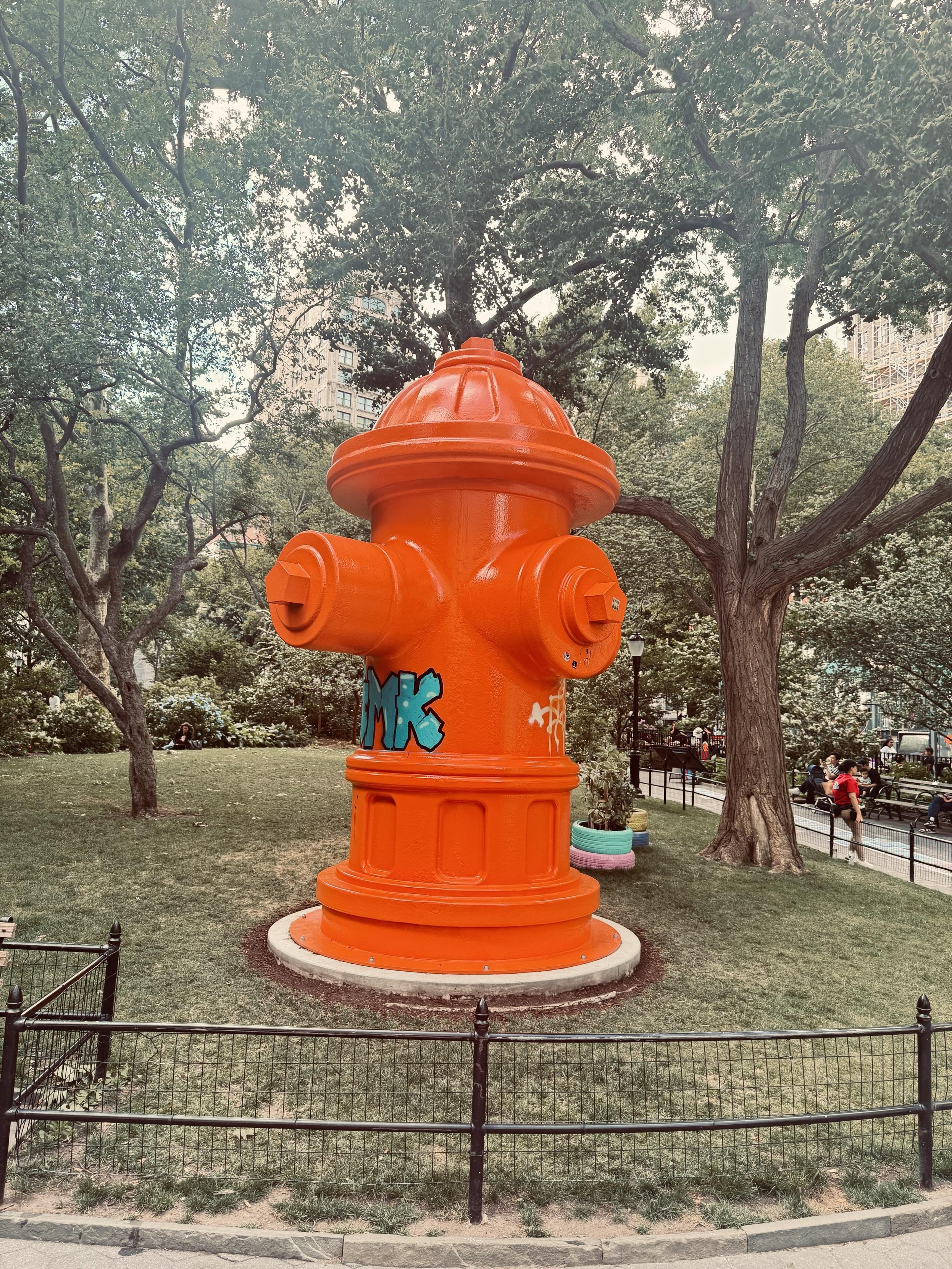 A color photo taken in Madison Square Park. An artist has put up a giant statue of a bright orange fire hydrant. A little bit of graffiti can be seen in blue on the left side and white on the right side. That is part of the art piece. Behind it are some painted tires in blue and pink with plants inside that are also part of the art. There is a low black railing in the front of the photo and two big trees on either side with more greenery in the back. The grass looks a little brownish from being dry at the beginning of the summer.